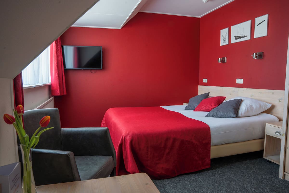 Een hotelkamer met een rode muur, een bed met kussens, een aan de muur bevestigde televisie, een stoel en een tafel.