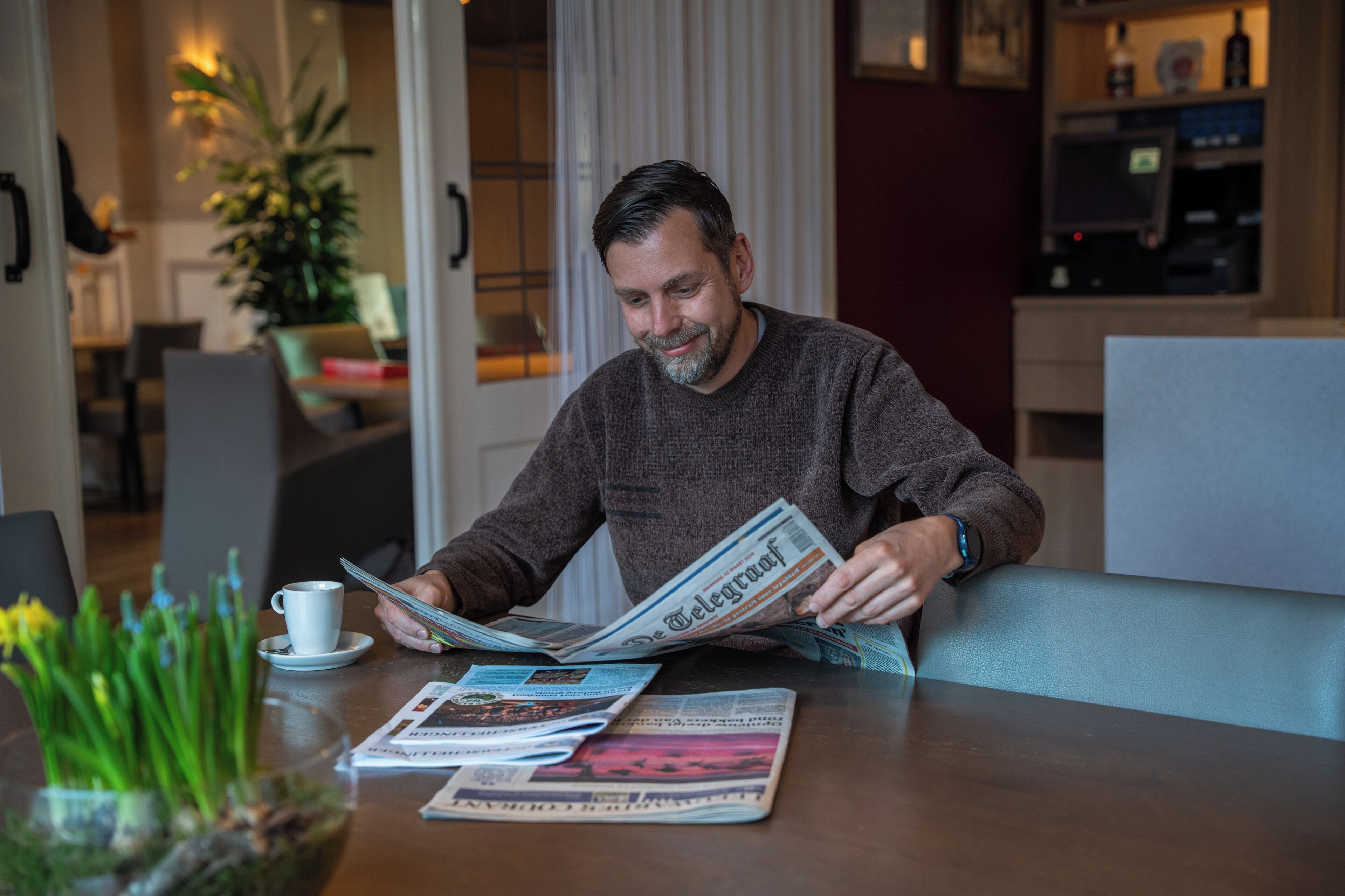 A man reading a newspaper at a table with a cup and saucer, a vase with green plants, and a magazine.