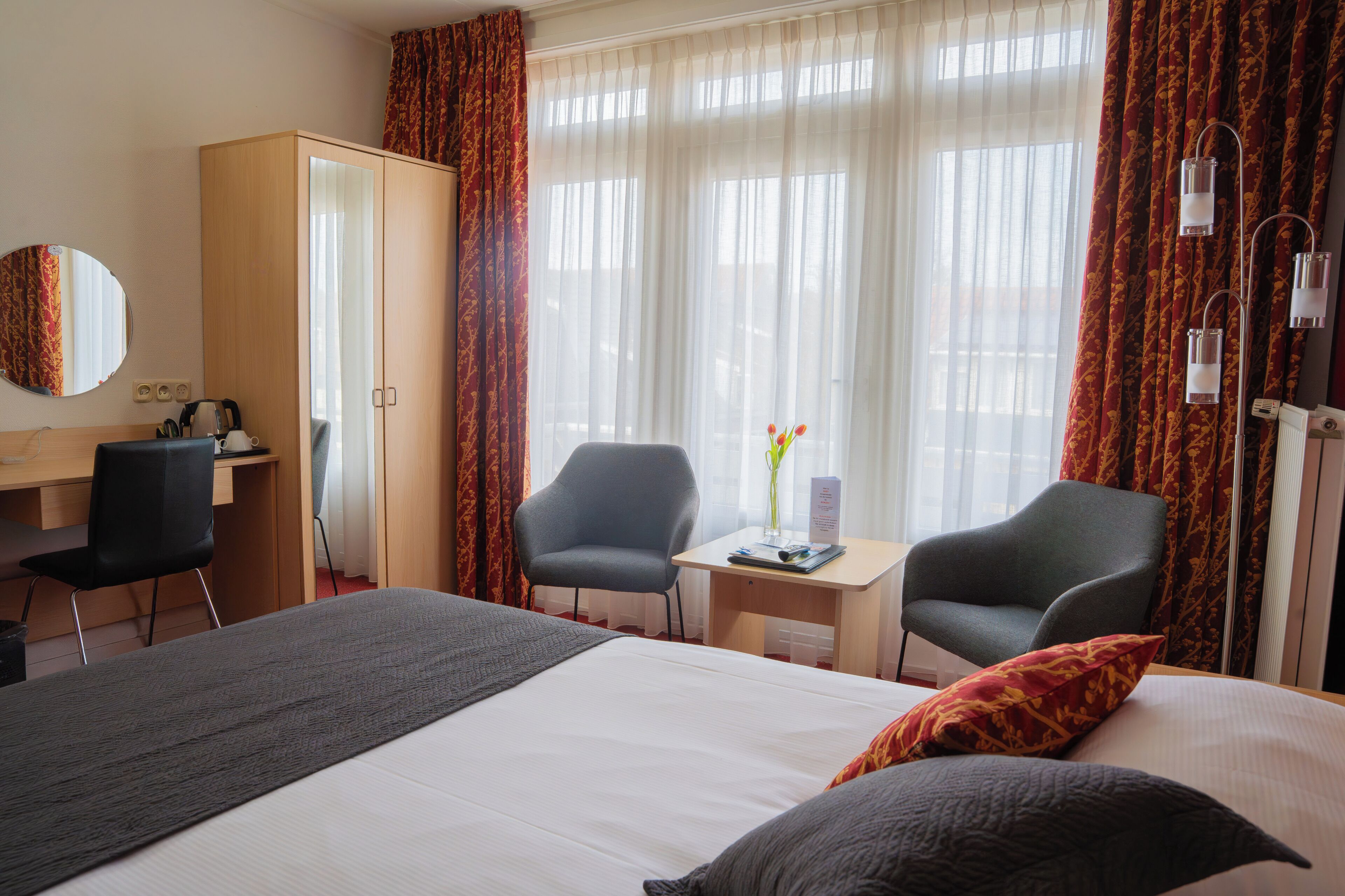 Habitación de hotel con cama, dos sillones, escritorio, armario y ventana con cortinas.