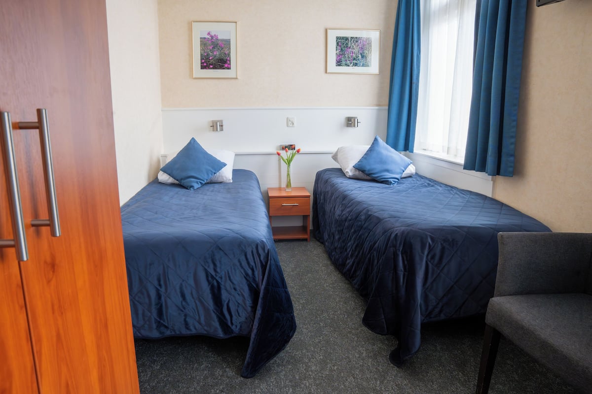 Een hotelkamer met twee bedden, een houten deur, een kleine tafel met een vaas en een raam met gordijnen.