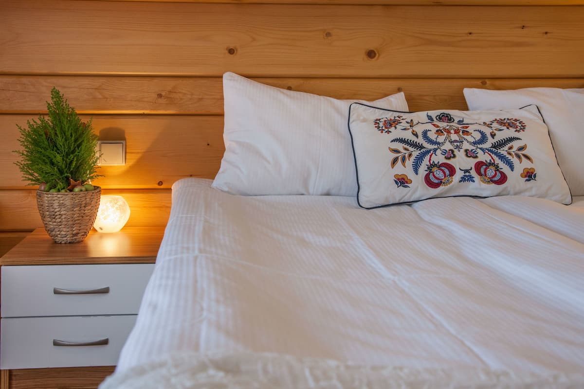 Un lit bien fait, avec du linge de lit blanc et un coussin décoratif, une lampe de chevet et une petite plante en pot sur une table de chevet en bois.