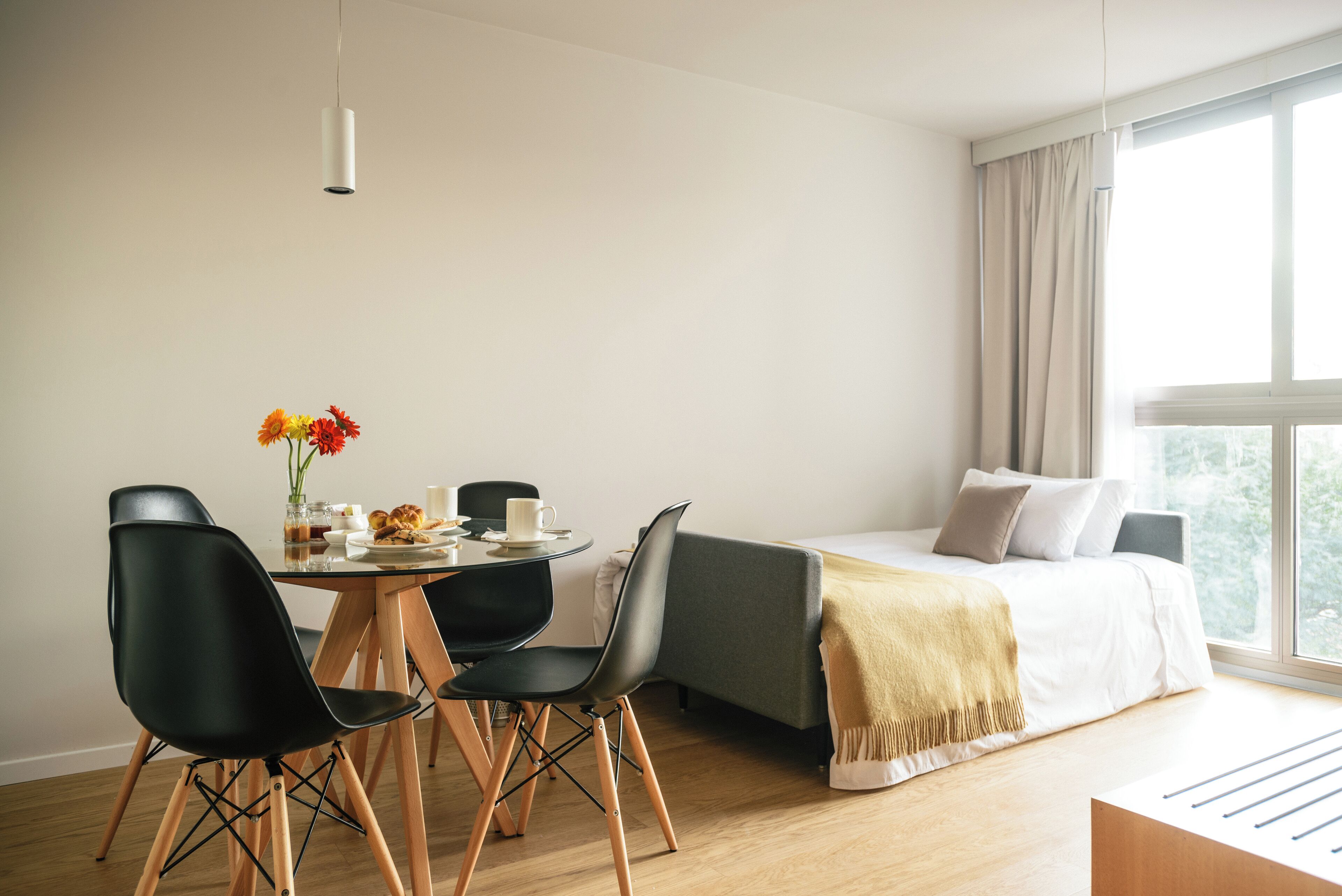 A modern room with a bed, a dining table with chairs, a vase of flowers, and a window with curtains.
