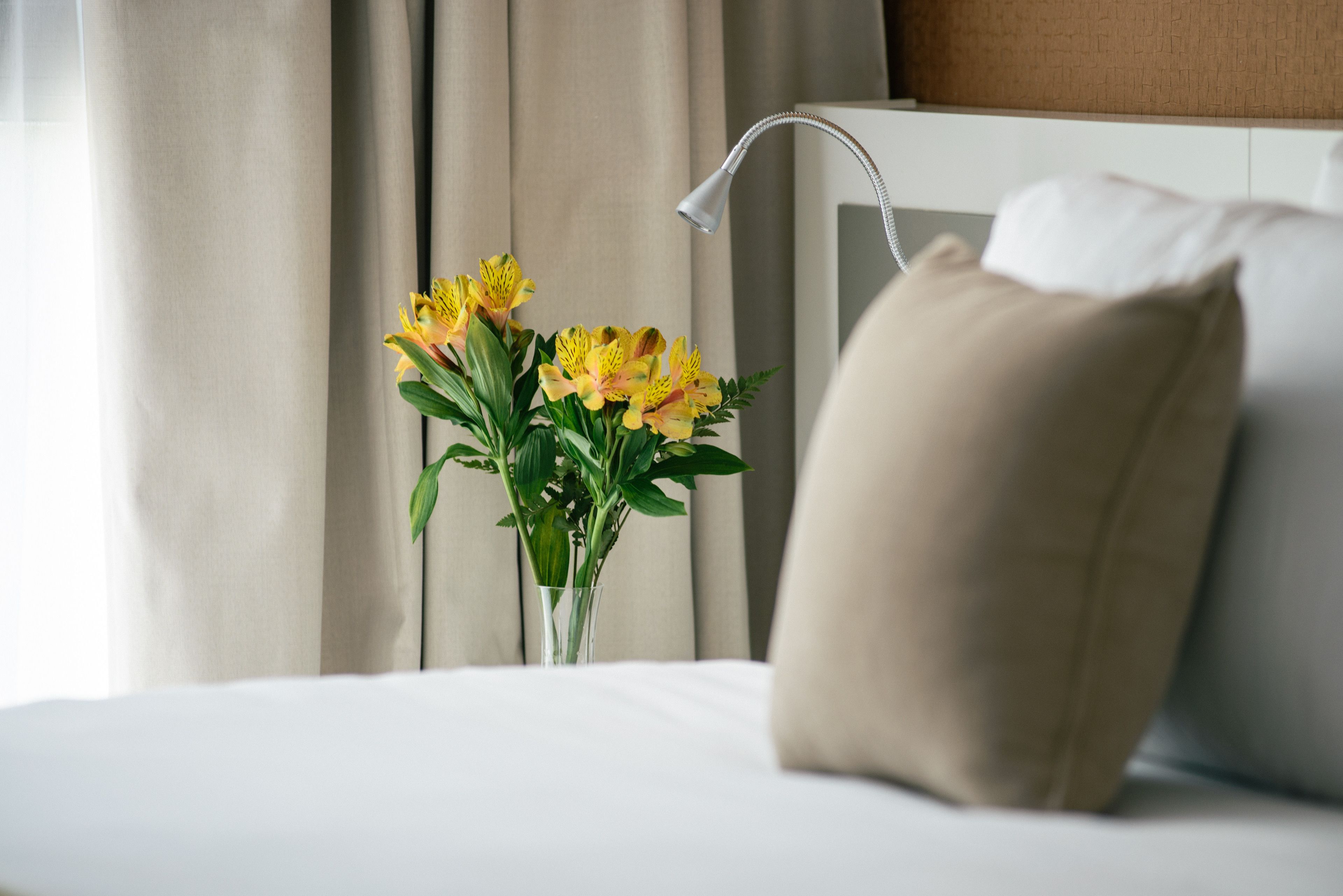Un lit avec un oreiller beige, une table de chevet blanche et un vase rempli de fleurs jaunes.