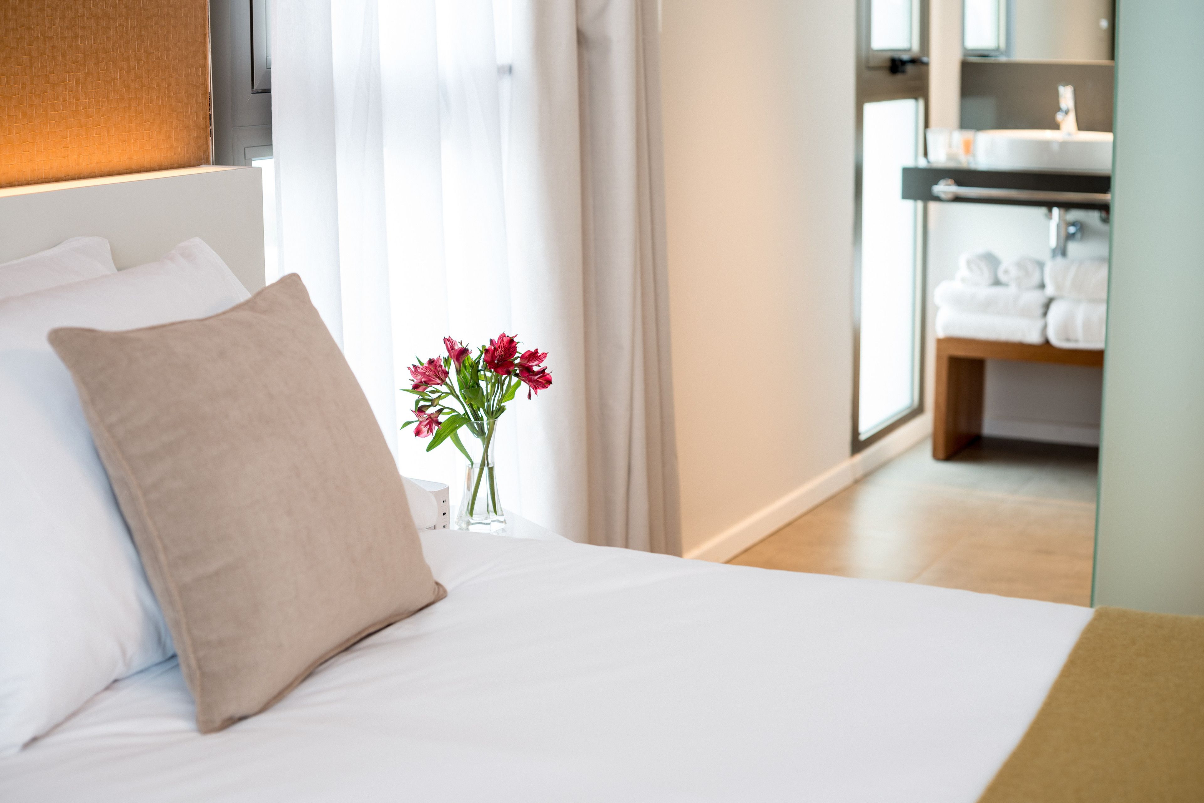 Una habitación de hotel con una cama, un florero con flores y un baño visible al fondo.