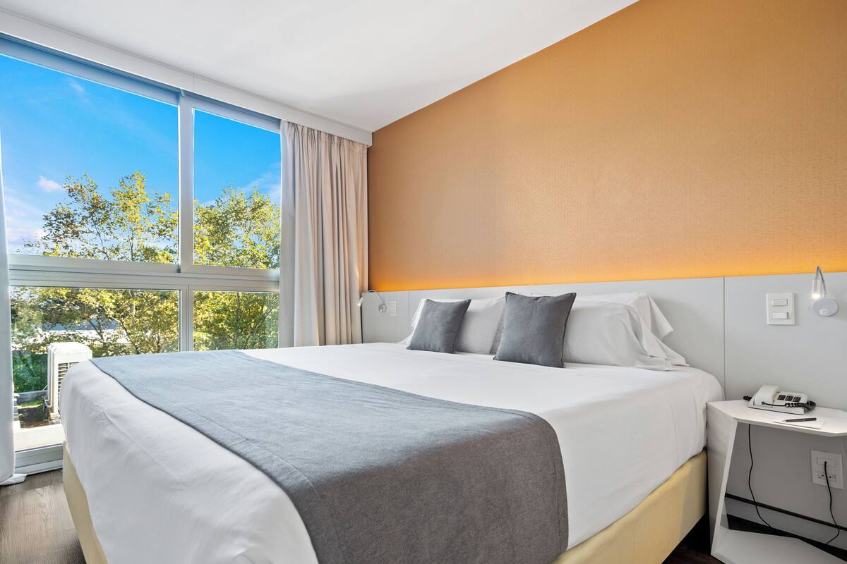 A modern hotel room with a large bed, a bedside table with a phone, and a view of trees through the window.