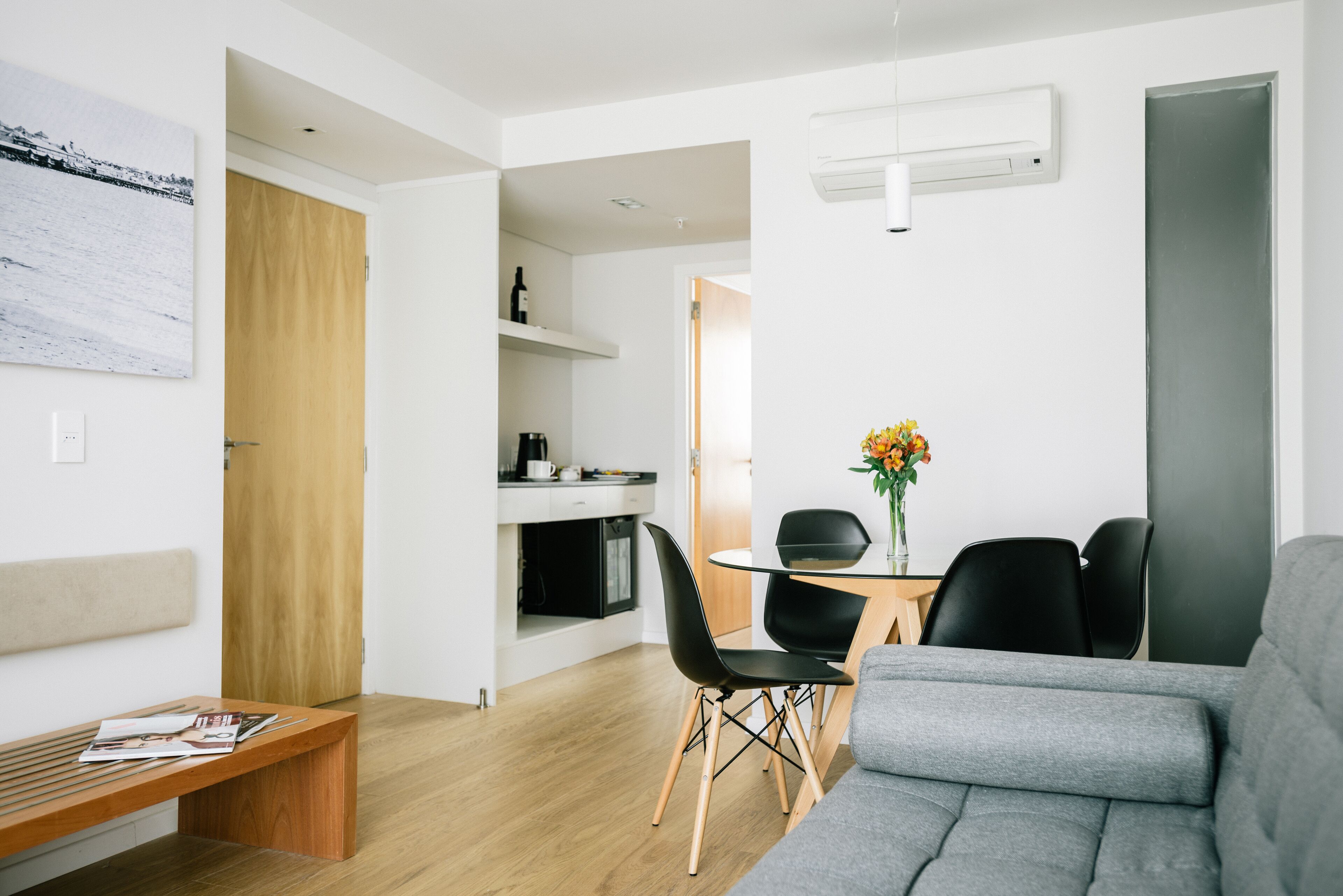 Uma sala de estar moderna com um sofá cinzento, um banco de madeira e uma área de jantar com mesa de vidro e cadeiras pretas.