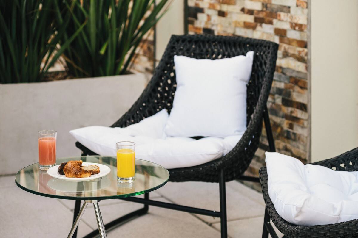 A glass table with a plate of food and two glasses of juice, a wicker chair with a white cushion, and a potted plant in the background.