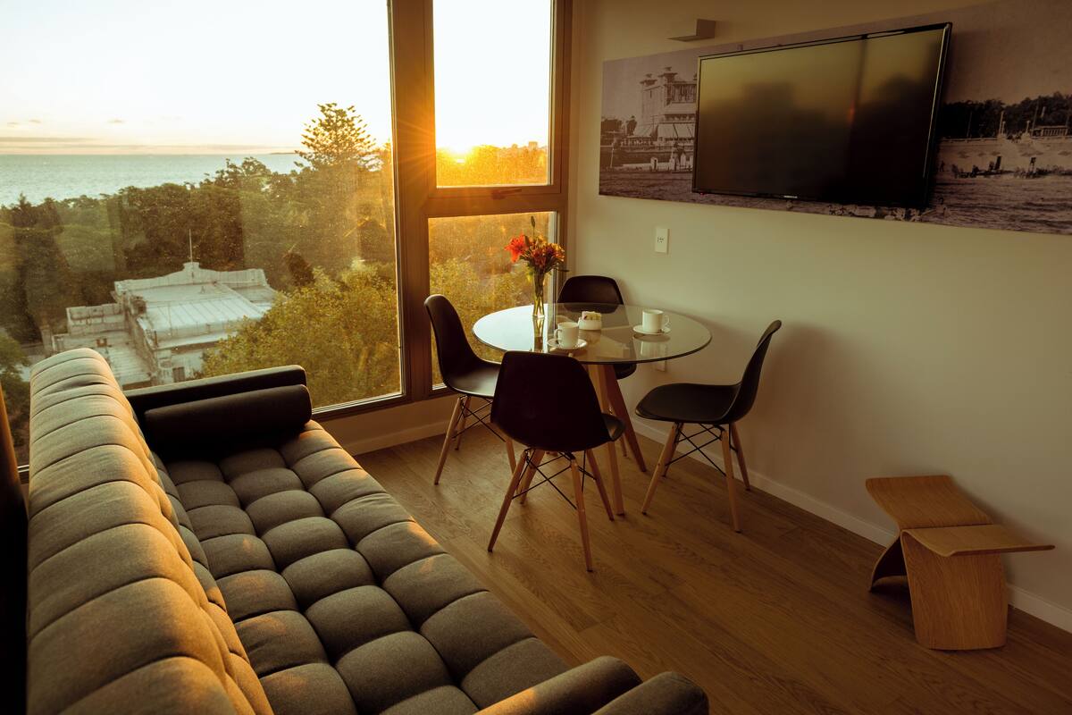 A modern living room with a large window, a sofa, a dining table with chairs, and a TV mounted on the wall.