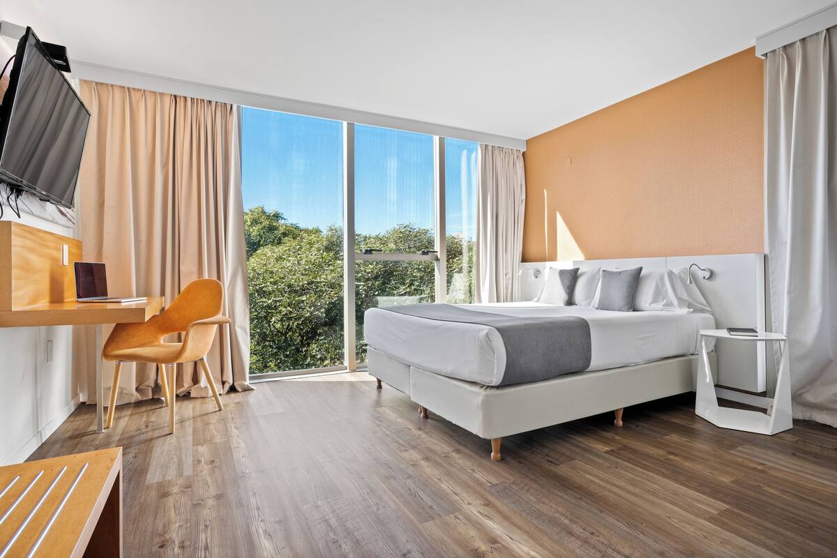 A modern hotel room with a large bed, a desk with a chair, a TV, and a view of greenery through large windows.