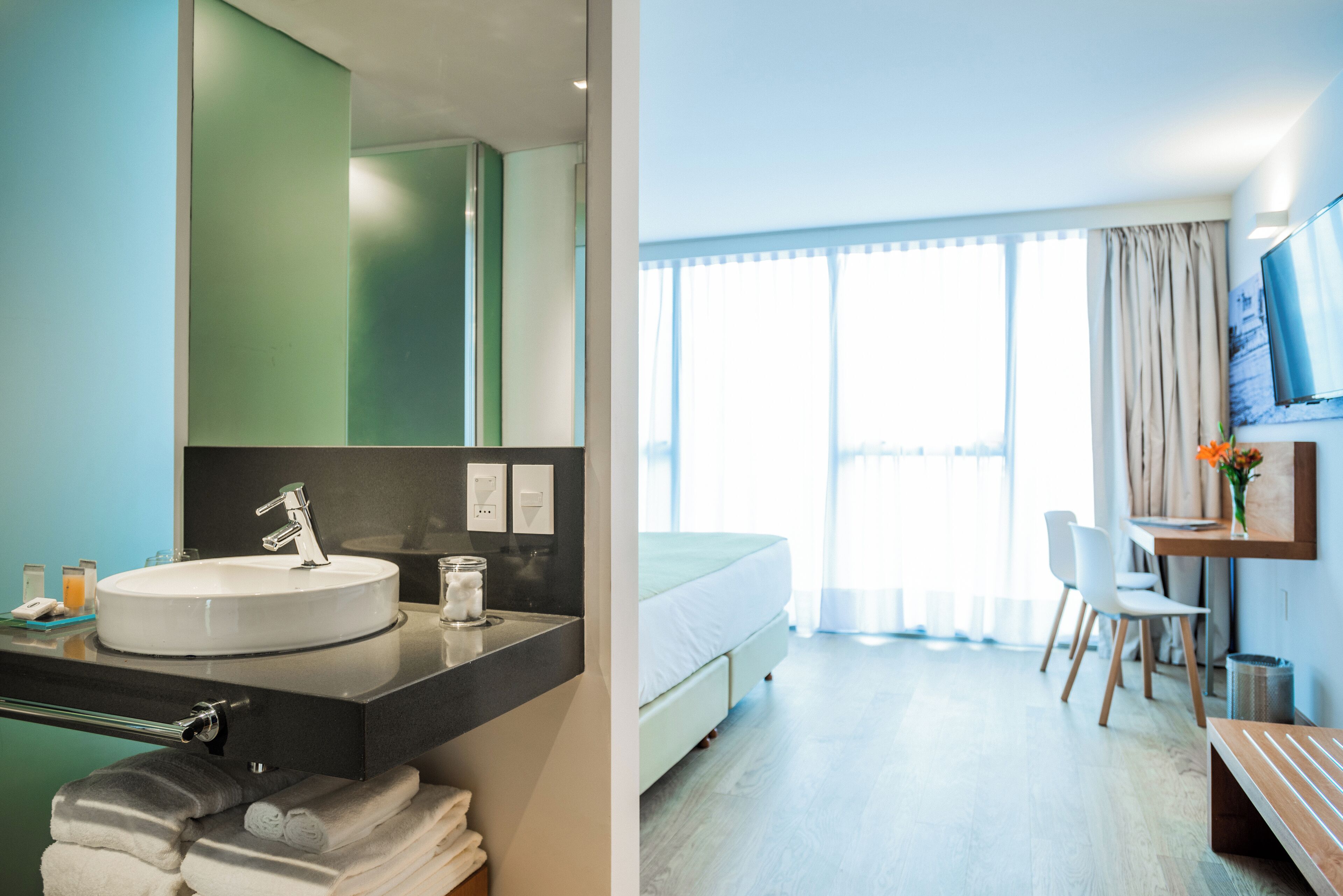 A hotel room with a large bed, a bathroom with a sink and mirror, a desk with a chair, and a TV on the wall.