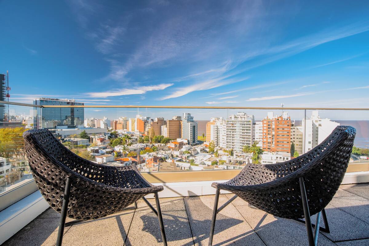 A balcony with two wicker chairs overlooking a cityscape with buildings and a body of water.