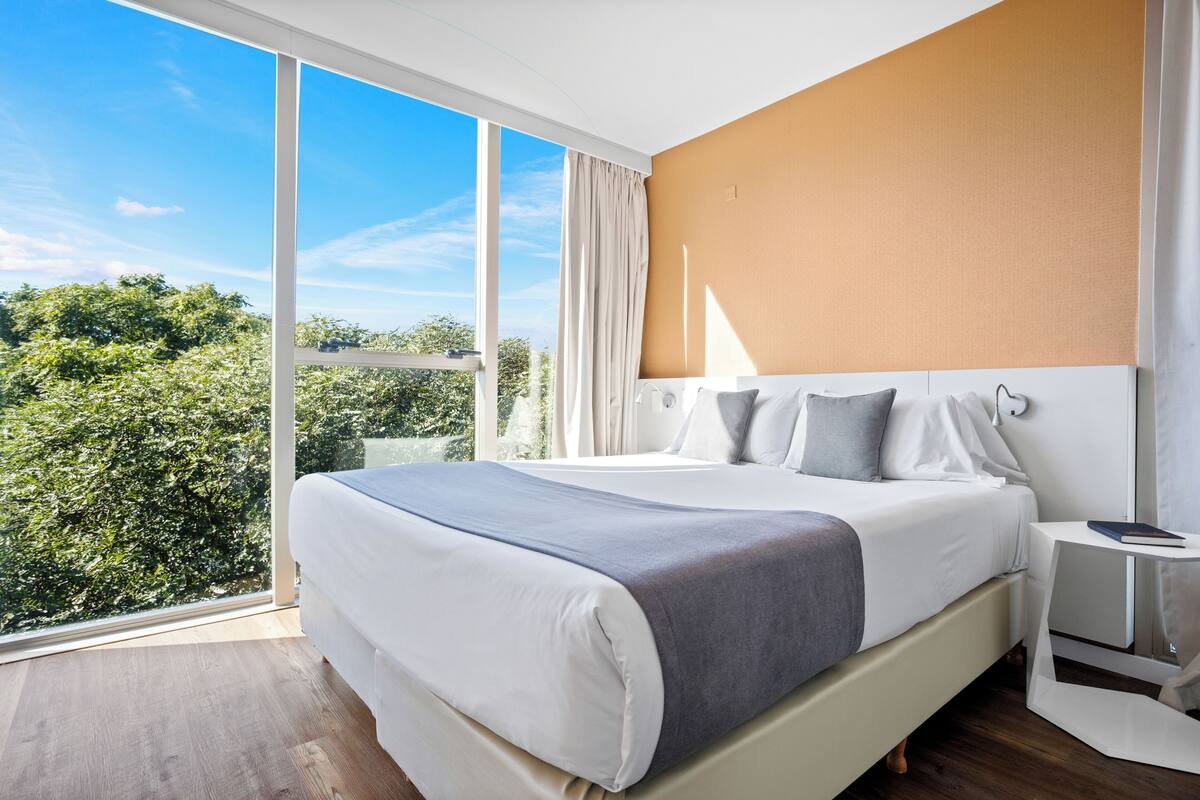 A modern hotel room with a large bed, a bedside table, and a view of greenery through floor-to-ceiling windows.