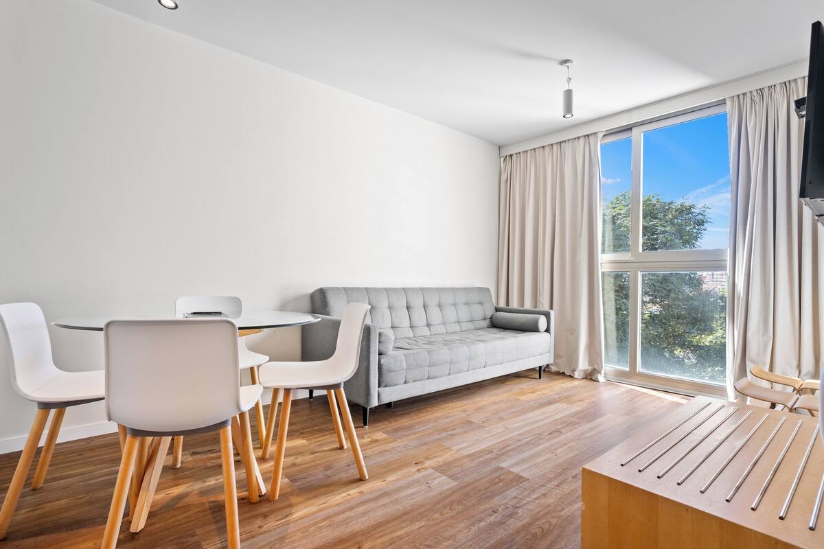 A modern living room with a grey sofa, white chairs, a wooden coffee table, and a large window with curtains.