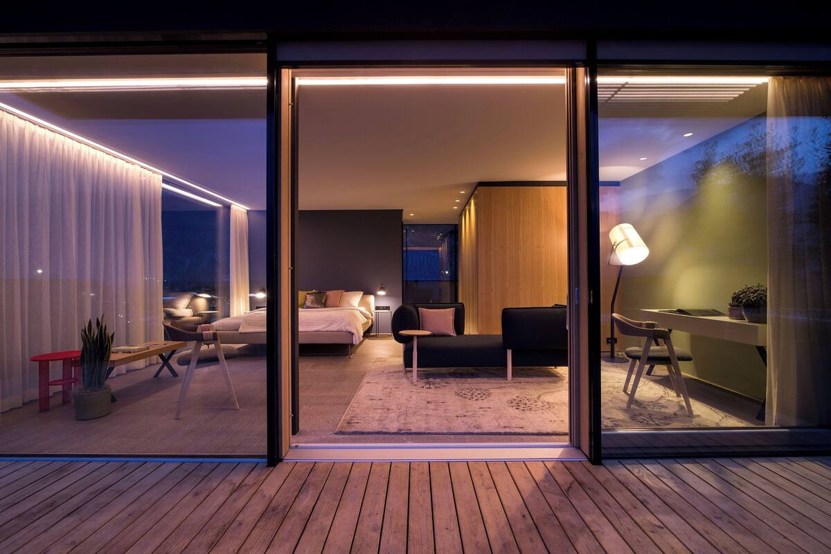 A modern hotel room with a wooden floor, a bed with white bedding, a black sofa, a small table, and a desk with a lamp.
