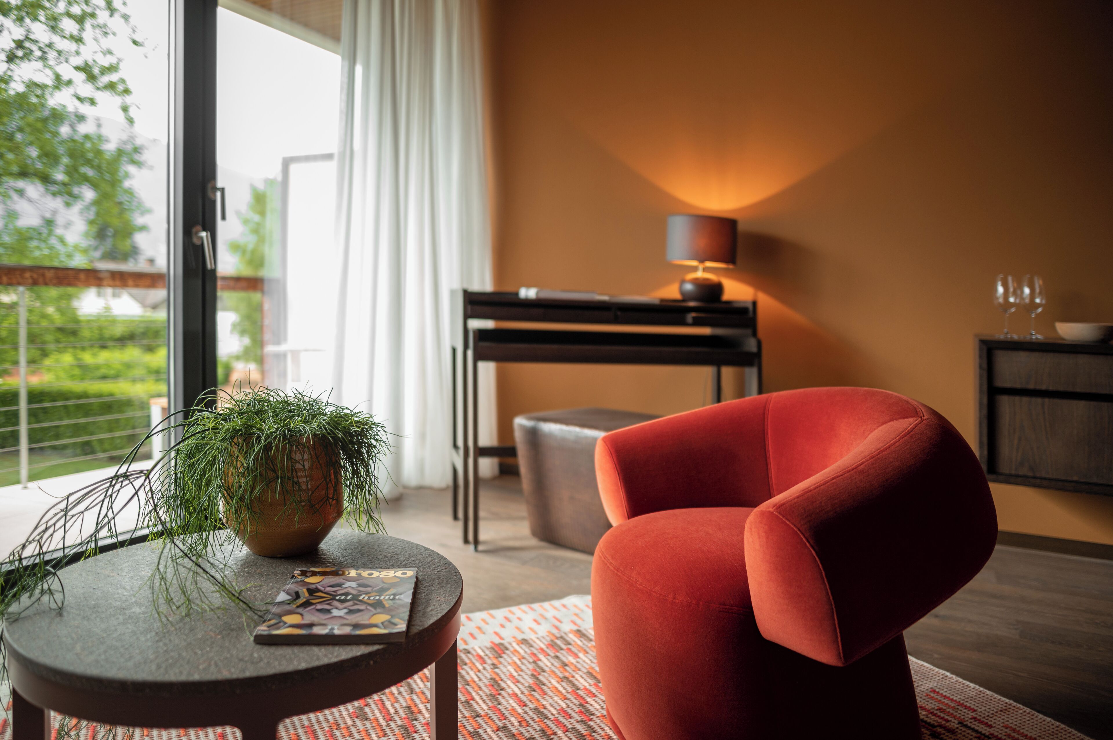 Una sala de estar moderna con un sillón rojo, una mesita redonda con una planta, una lámpara y vistas al exterior.