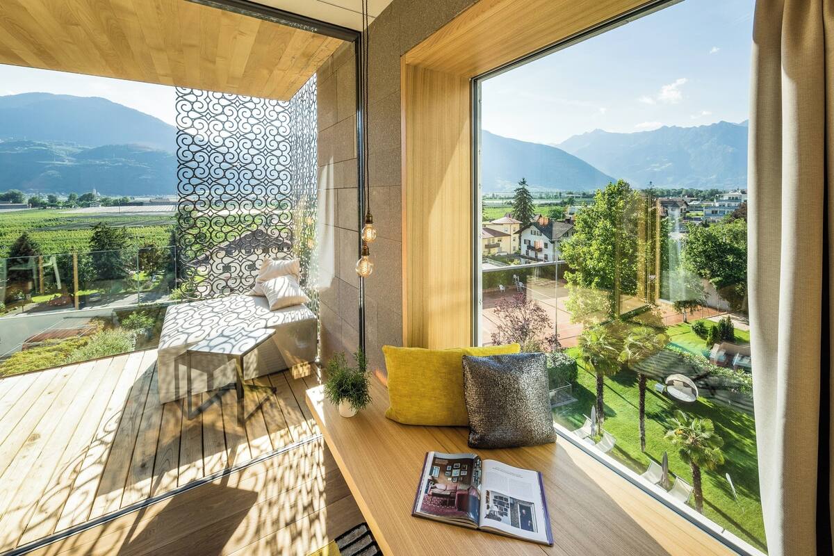 A modern room with a large window overlooking a scenic landscape, a wooden deck, and a cozy seating area with cushions.