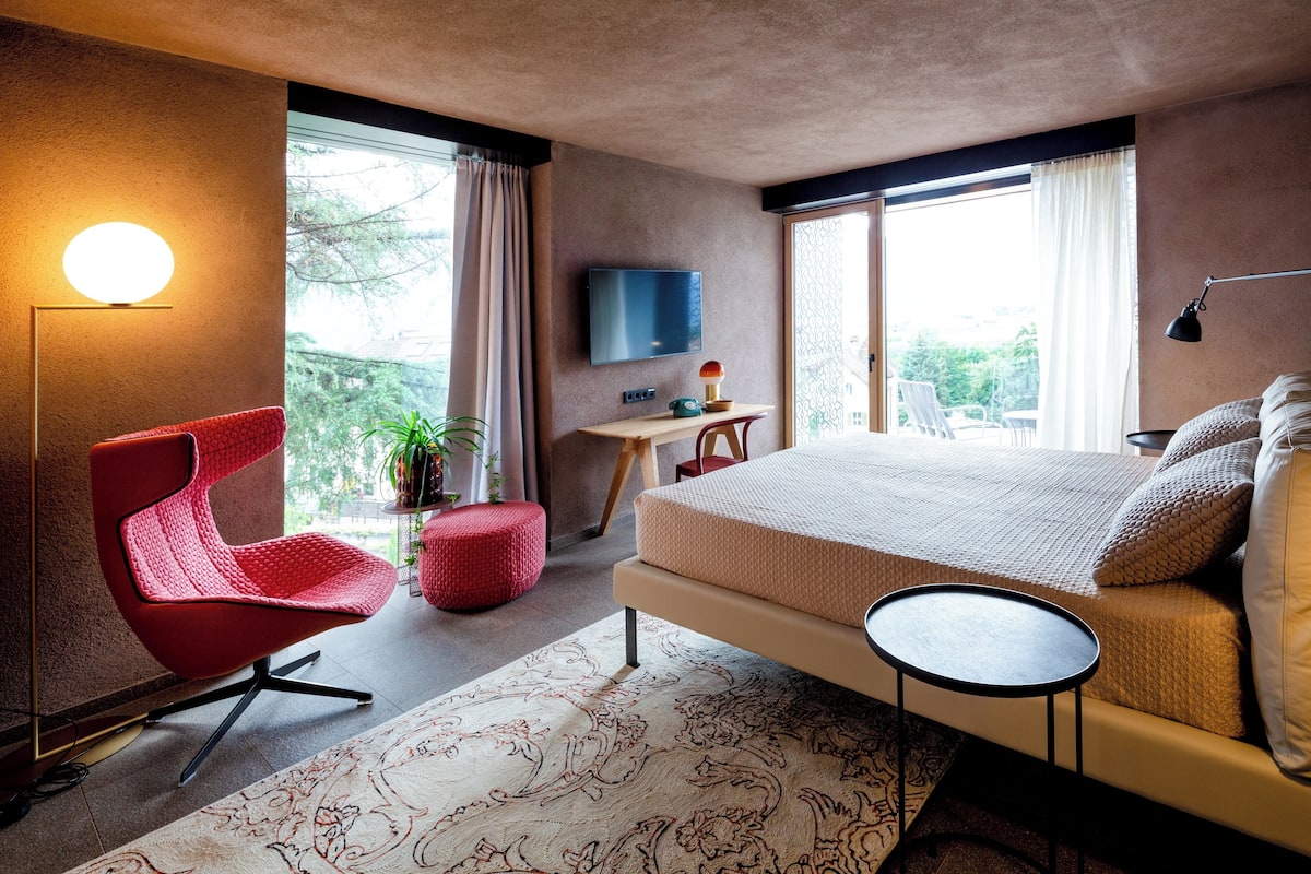 A modern bedroom with a large bed, a red chair, a small desk, and a TV on the wall.