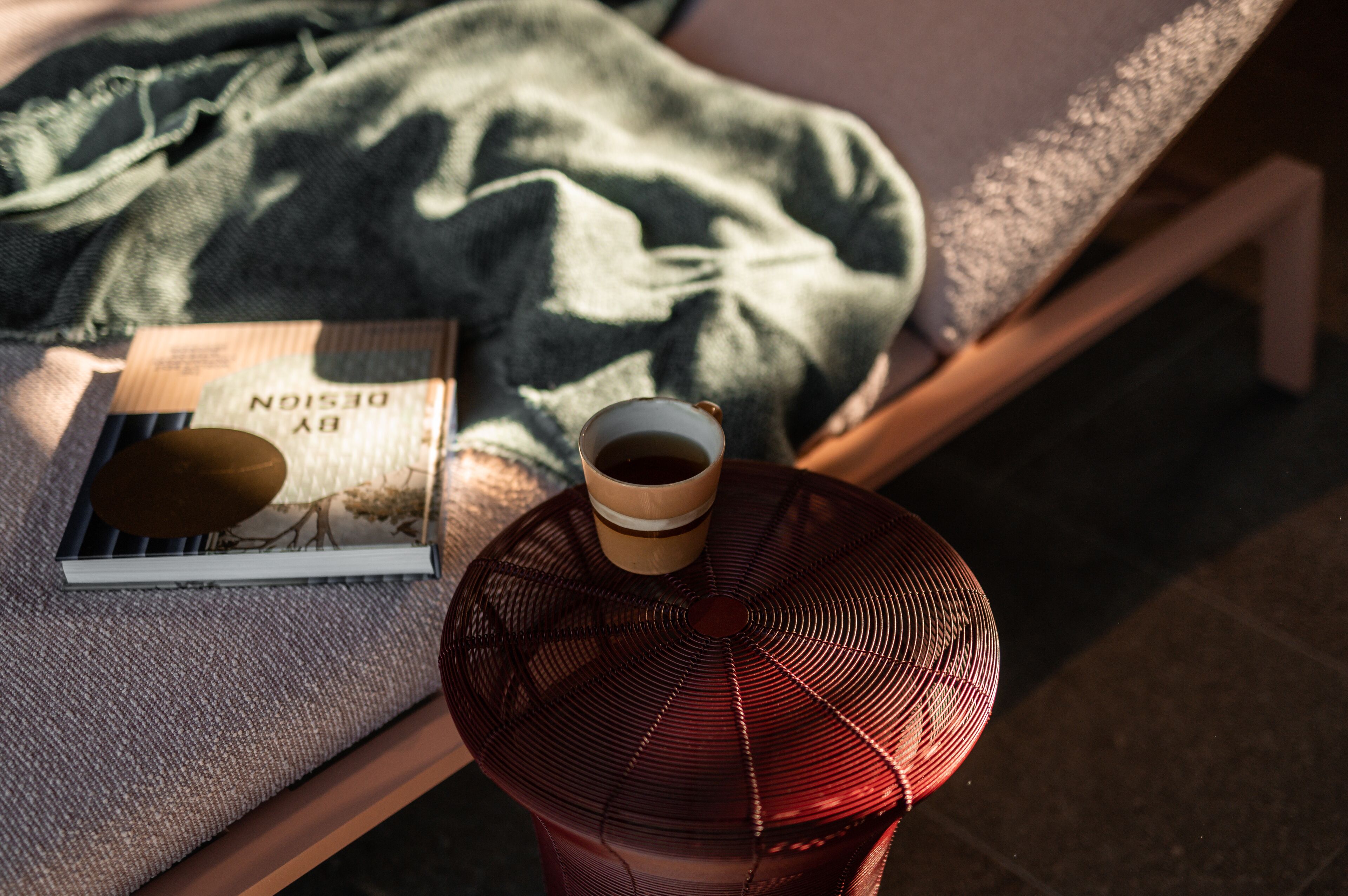 A bed with a green blanket, a book, a cup of coffee, and a red fan on a wooden table.