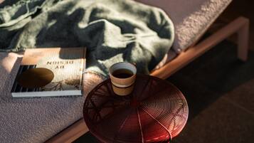 A bed with a green blanket, a book, a cup of coffee, and a red fan on a wooden table.