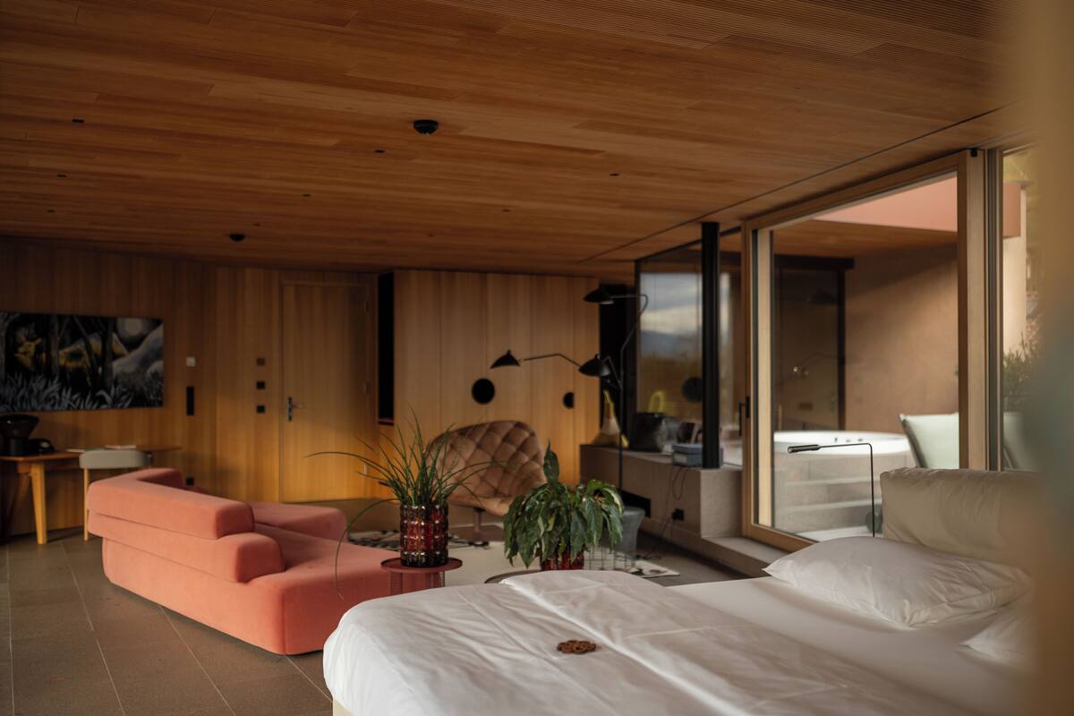 A modern bedroom with a large bed, a pink sofa, a wooden ceiling, and a large window.