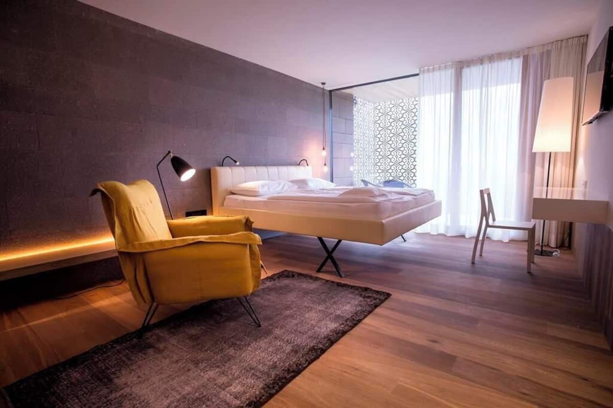 A modern hotel room with a bed, a yellow armchair, a desk, and a large window with curtains.