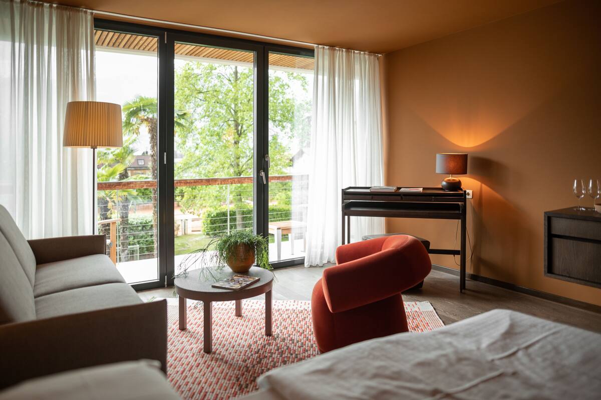 A hotel room with a sofa, a red chair, a desk, a lamp, and a view of greenery outside.