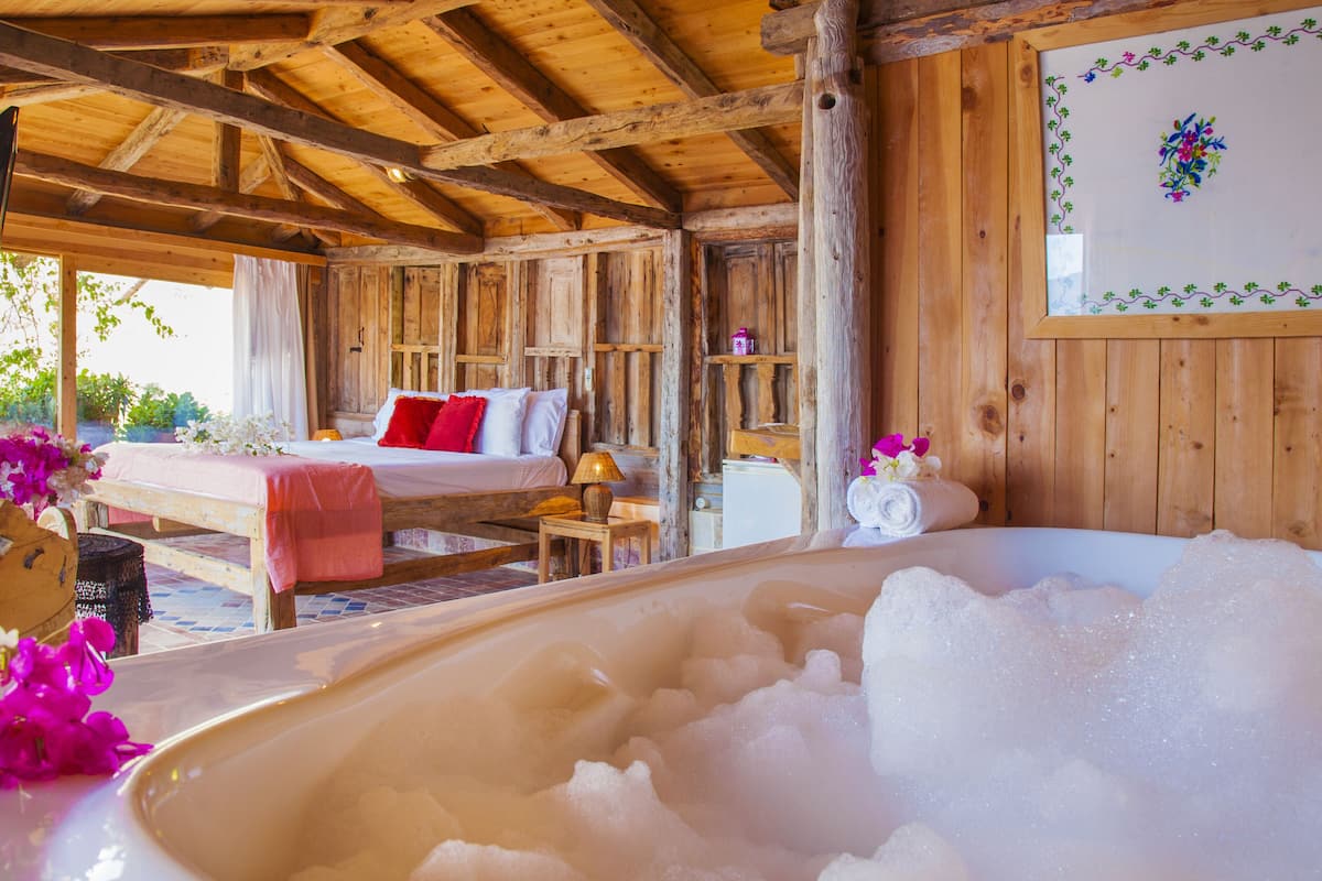 A wooden cabin with a bedroom, a bathroom with a bathtub filled with bubbles, and a wooden ceiling.