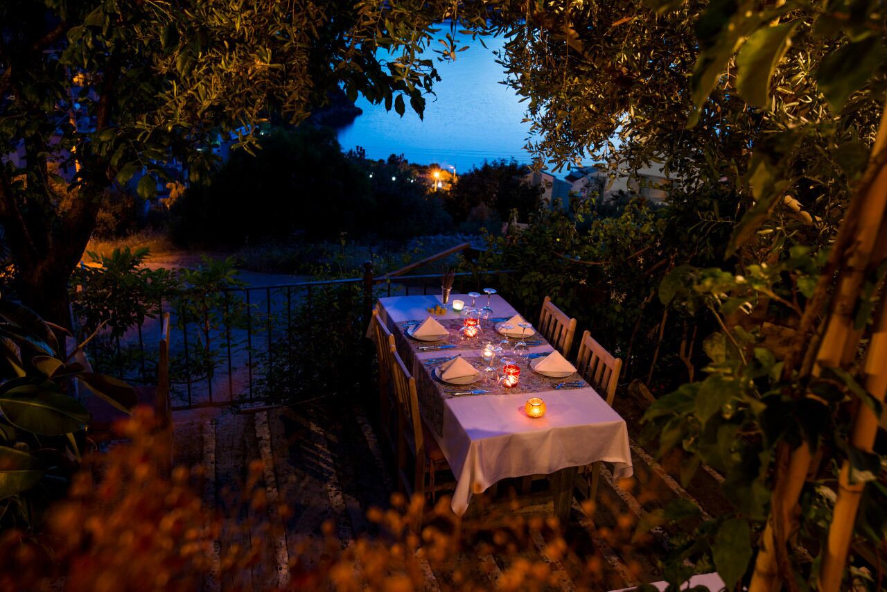 An outdoor dining table set up with candles, surrounded by foliage and overlooking a body of water.
