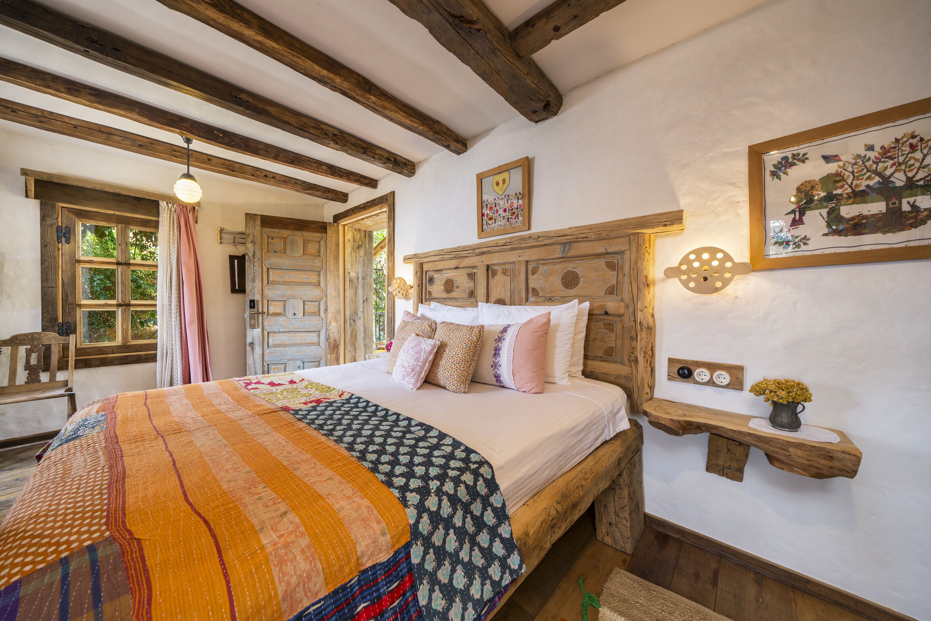 A cozy bedroom with a wooden bed, a colorful quilt, a wooden headboard, a wall-mounted shelf with a vase, and a window with curtains.