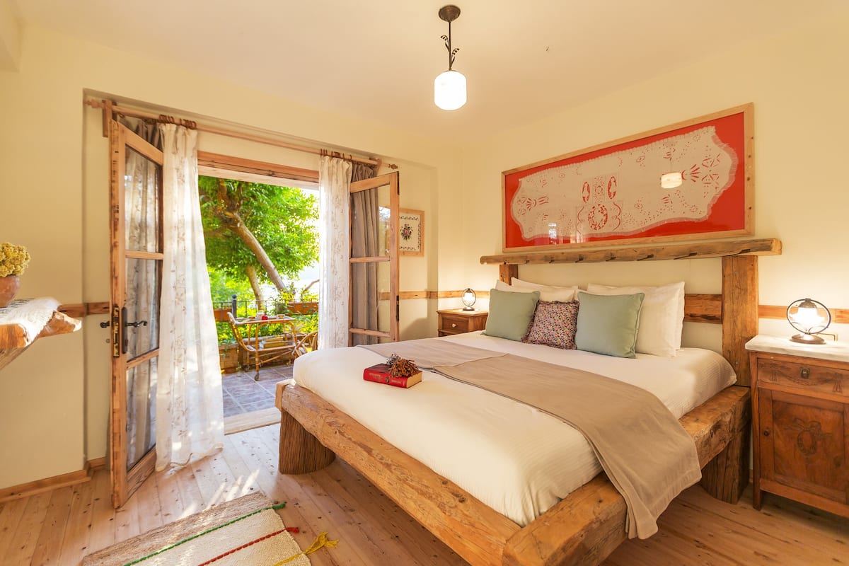 A wooden bed with a headboard, a bedside table, and a nightstand. There is a window with curtains and a view of greenery outside.