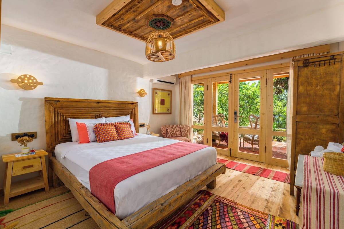 A wooden bed with a red blanket, a wooden nightstand, and a large window with a view of greenery.