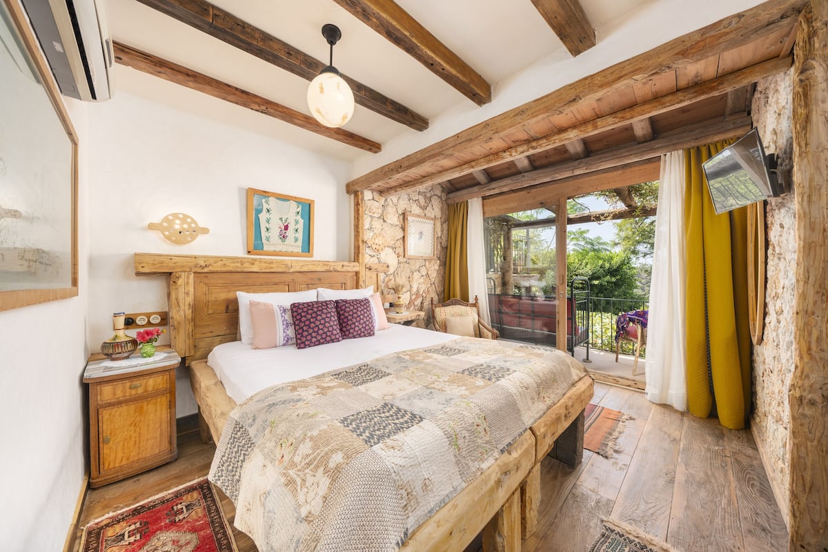A bedroom with a large bed, wooden beams, stone wall, and a view of greenery outside.
