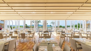 An outdoor dining area with tables and chairs, large windows, and a view of the sea. - Hotel Baia Di Conte (Alghero)