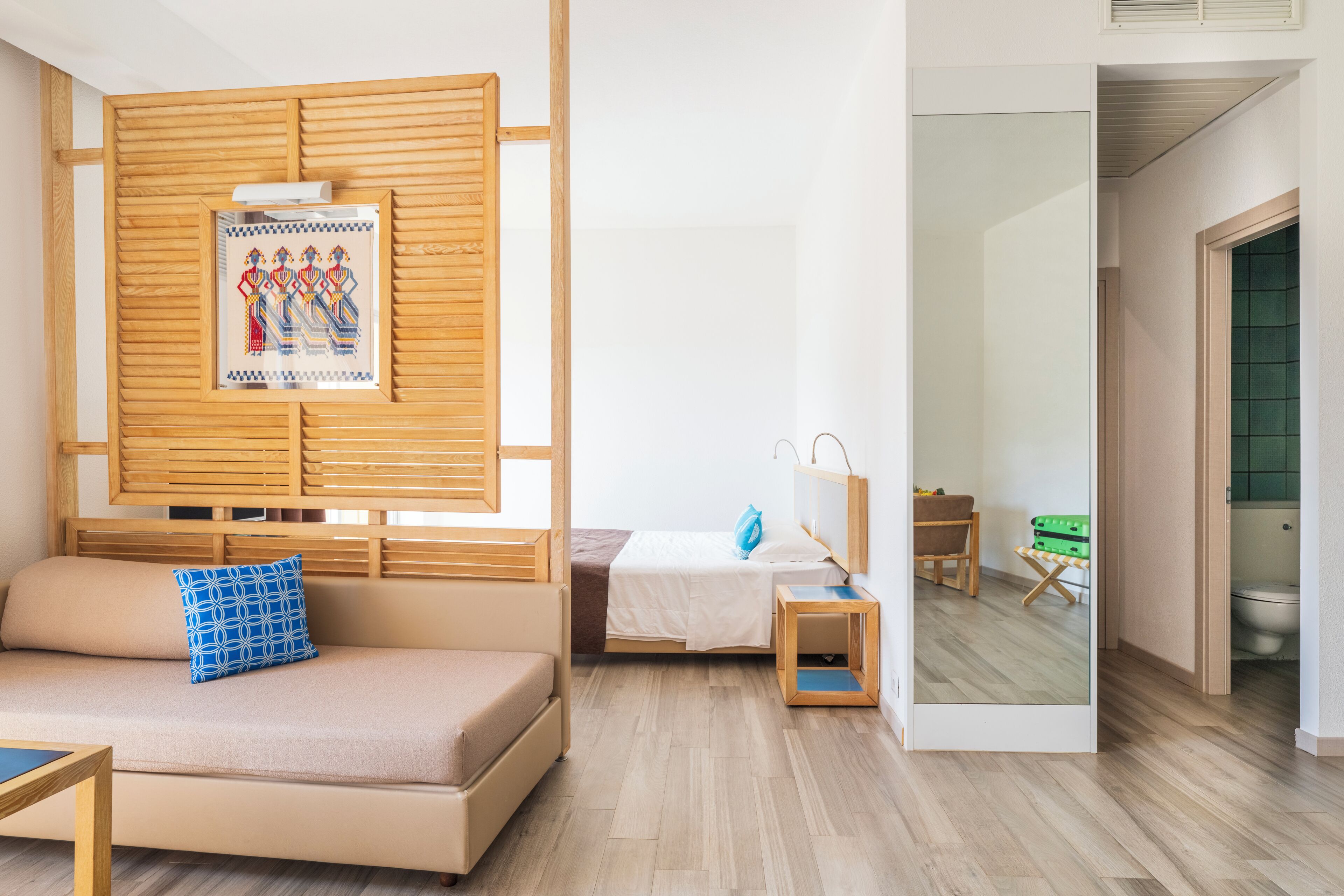 A modern hotel room with a sofa, a bed, a small table, a mirror, and a wooden partition with artwork.