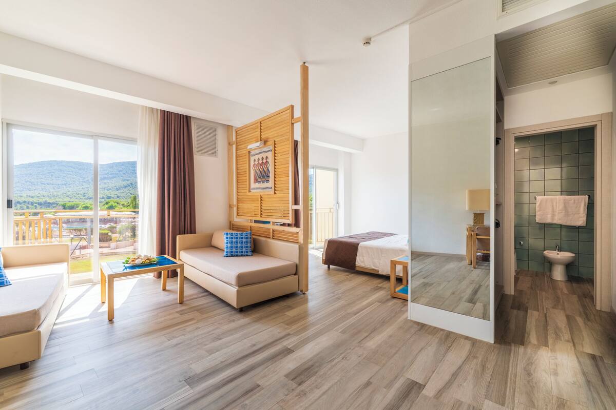 A hotel room with a sofa, a bed, a small table, a mirror, a bathroom with a sink and toilet, and a balcony with a view of the mountains.