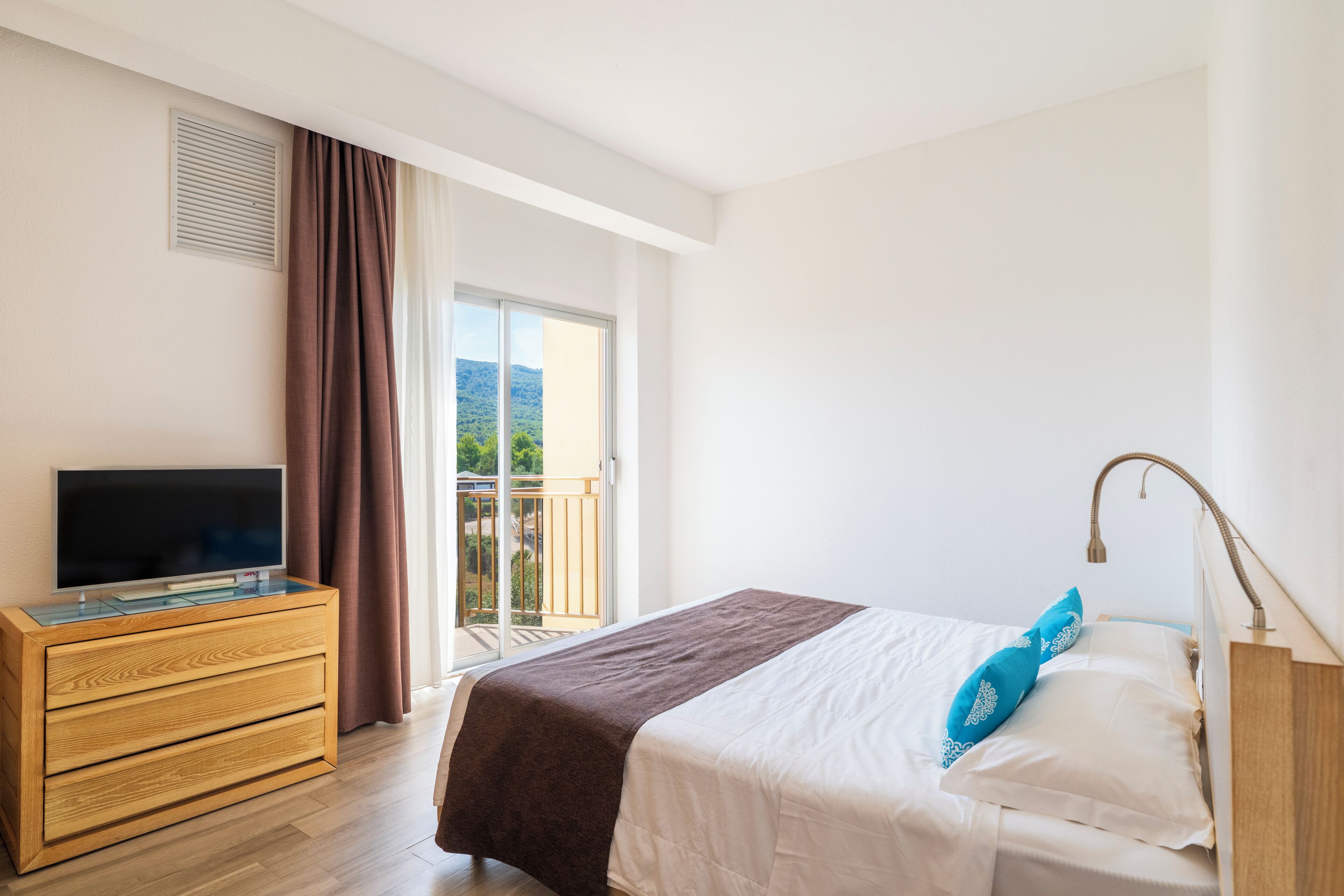 Habitación de hotel con cama, televisor en una cómoda de madera, balcón con vistas y ventana con cortinas.
