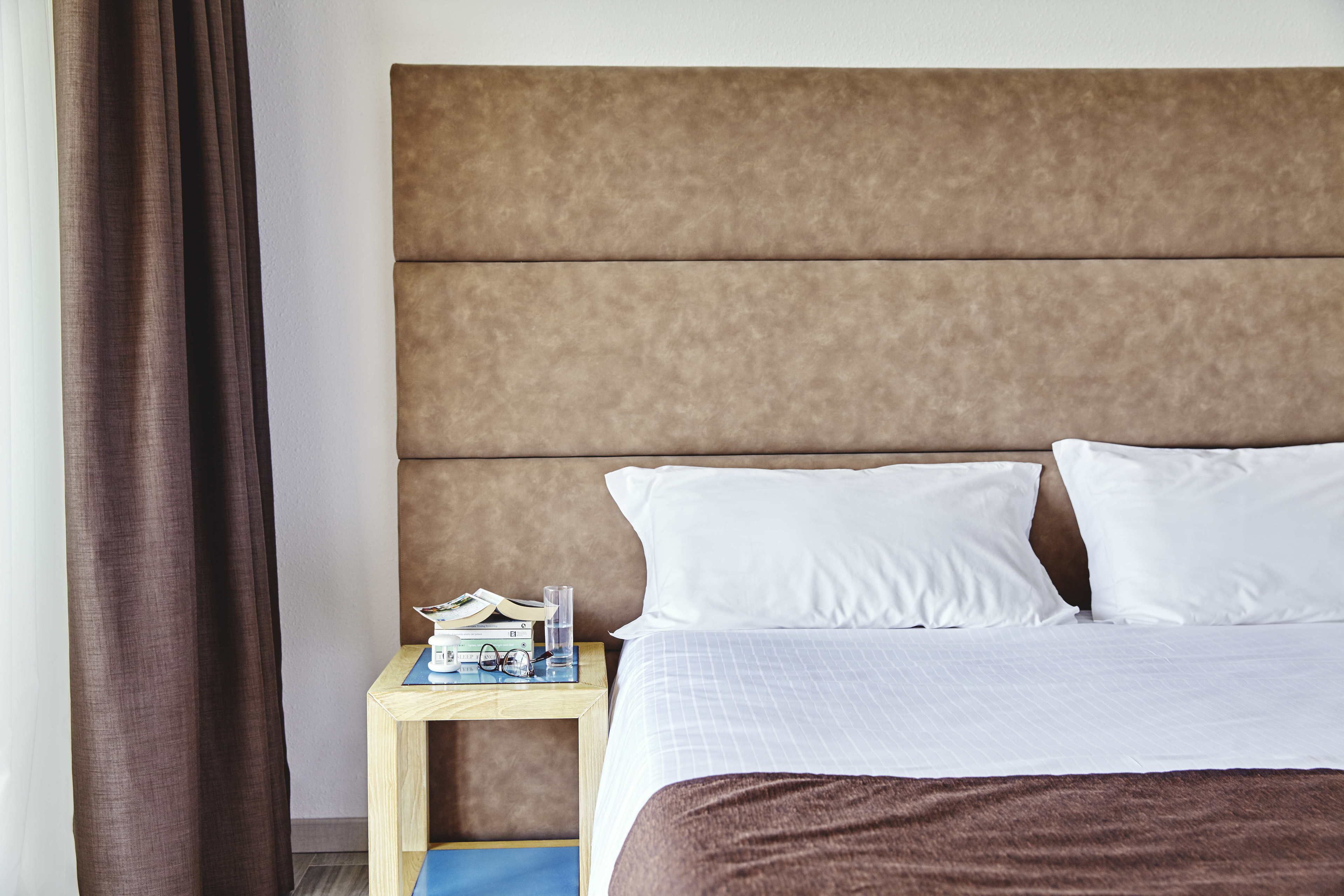 A neatly made bed with white linens and a brown headboard. A small wooden nightstand beside the bed holds a book, a glass of water, and a pair of glasses.