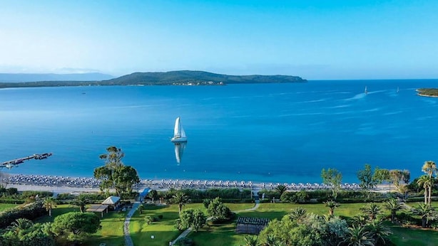 A coastal view with a sailboat on the water, a beach with people, and a park with greenery. - Hotel Baia Di Conte (Alghero)