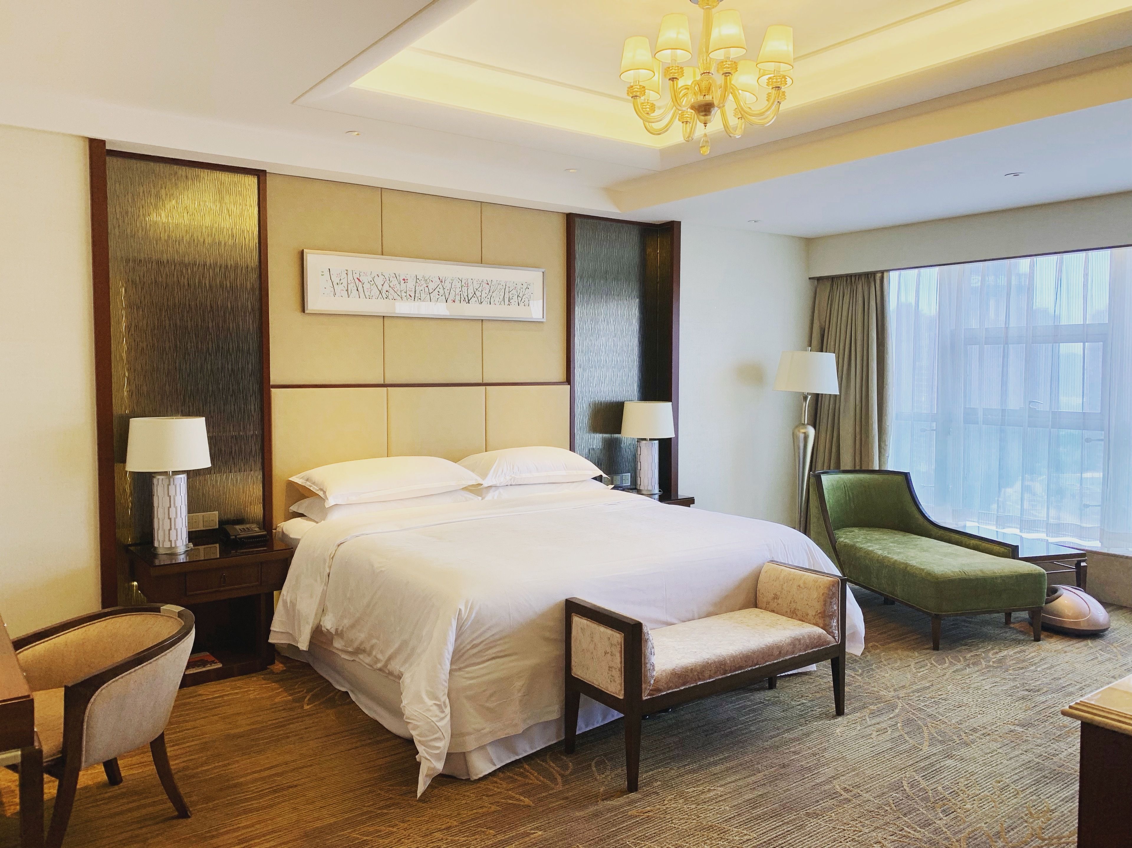 Habitación de hotel con una cama grande, dos mesitas de noche con lámparas, un sillón verde, un pequeño banco, un ventanal con cortinas y un candelabro.