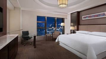 Una habitación de hotel con una cama grande, un escritorio, una silla, una mesita, una lámpara y vistas a la ciudad de noche.