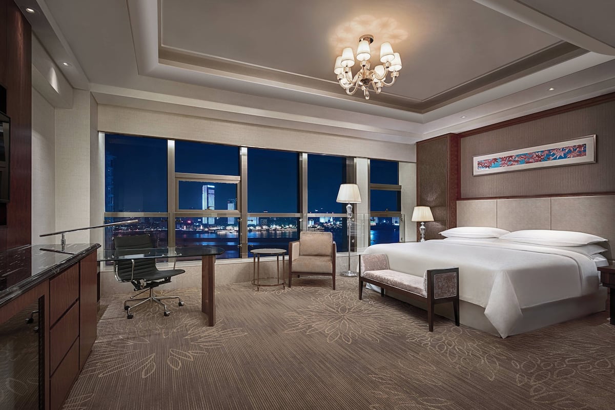 Habitación de hotel con una cama grande, un escritorio con silla, una mesita y vistas a la ciudad de noche.