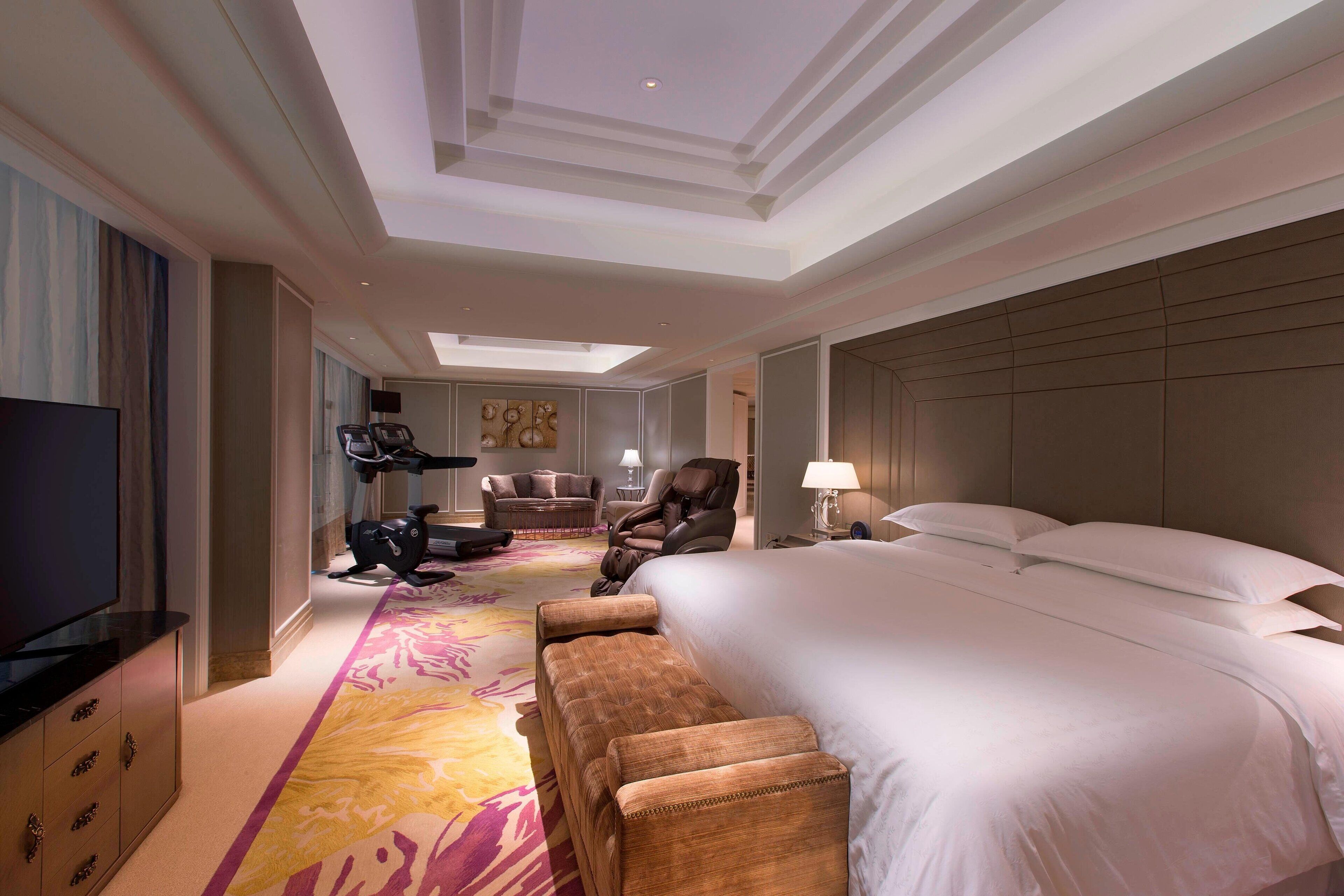 A hotel room with a large bed, a TV, exercise equipment, and a colorful carpet.