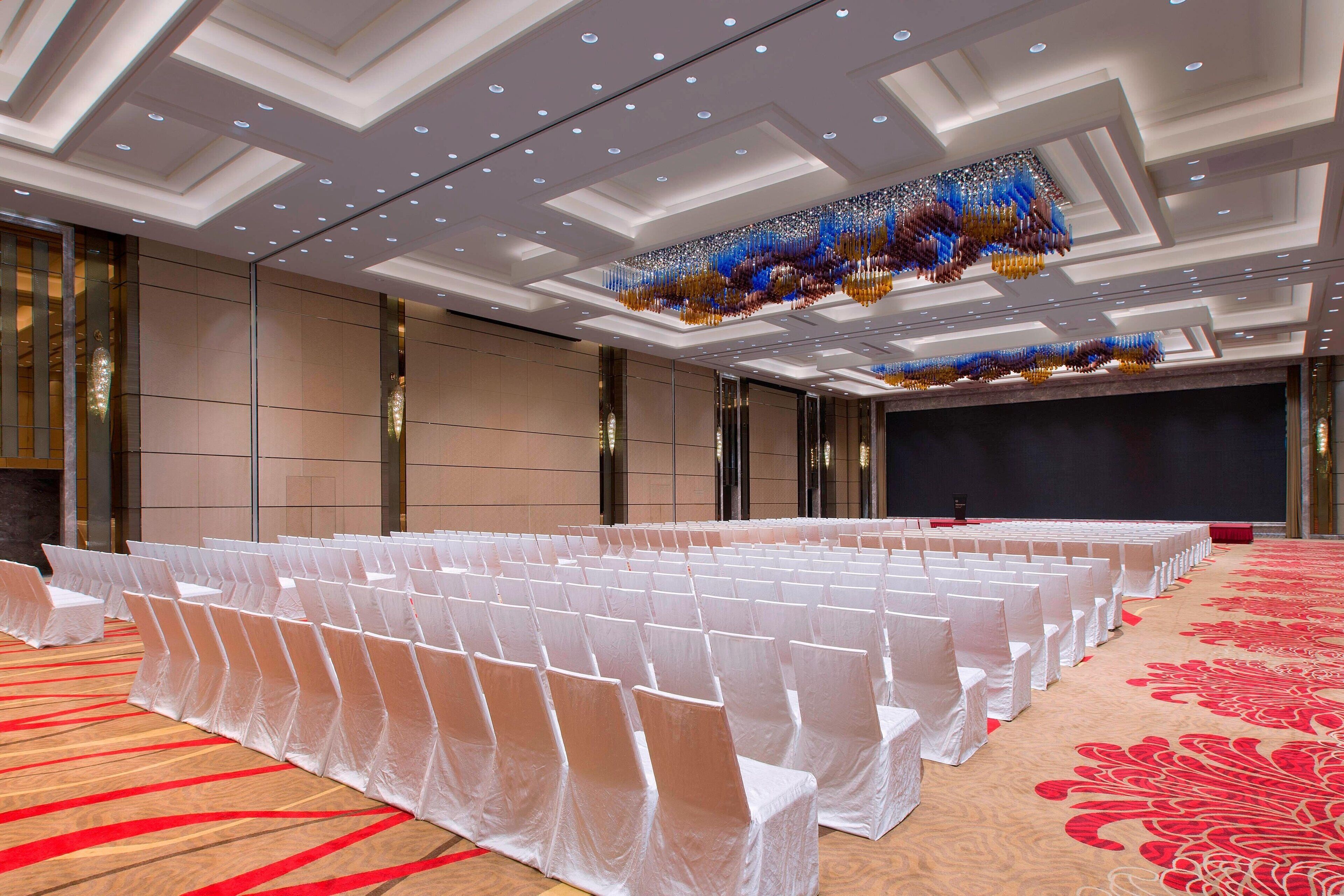 Un salón de conferencias con hileras de sillas blancas, una alfombra roja y una lámpara de araña moderna.