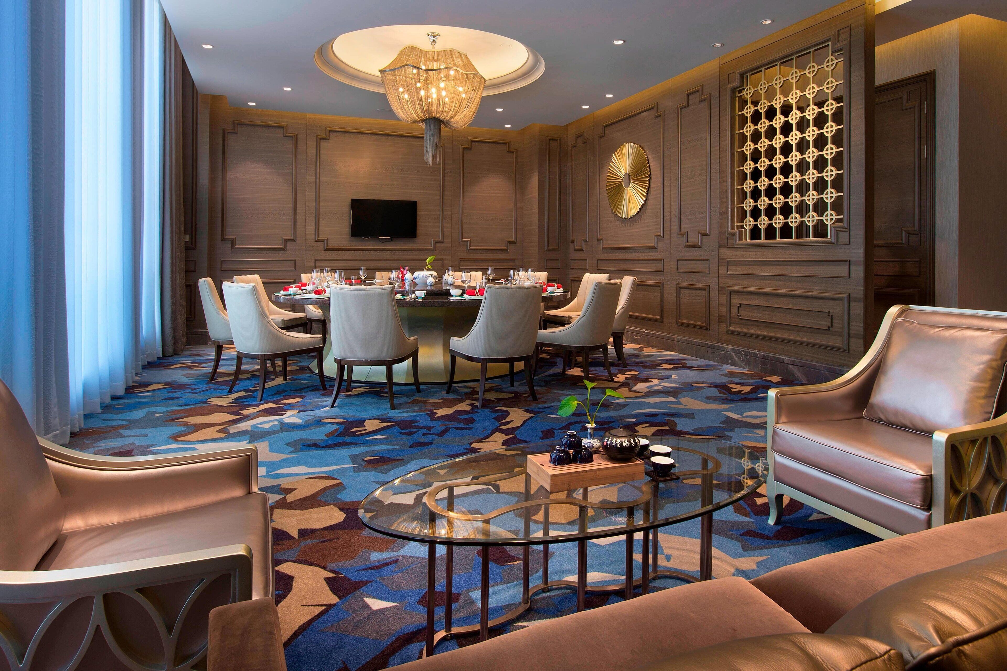 Una habitación de hotel moderna con zona de comedor, una mesa redonda, asientos cómodos y una lámpara decorativa.