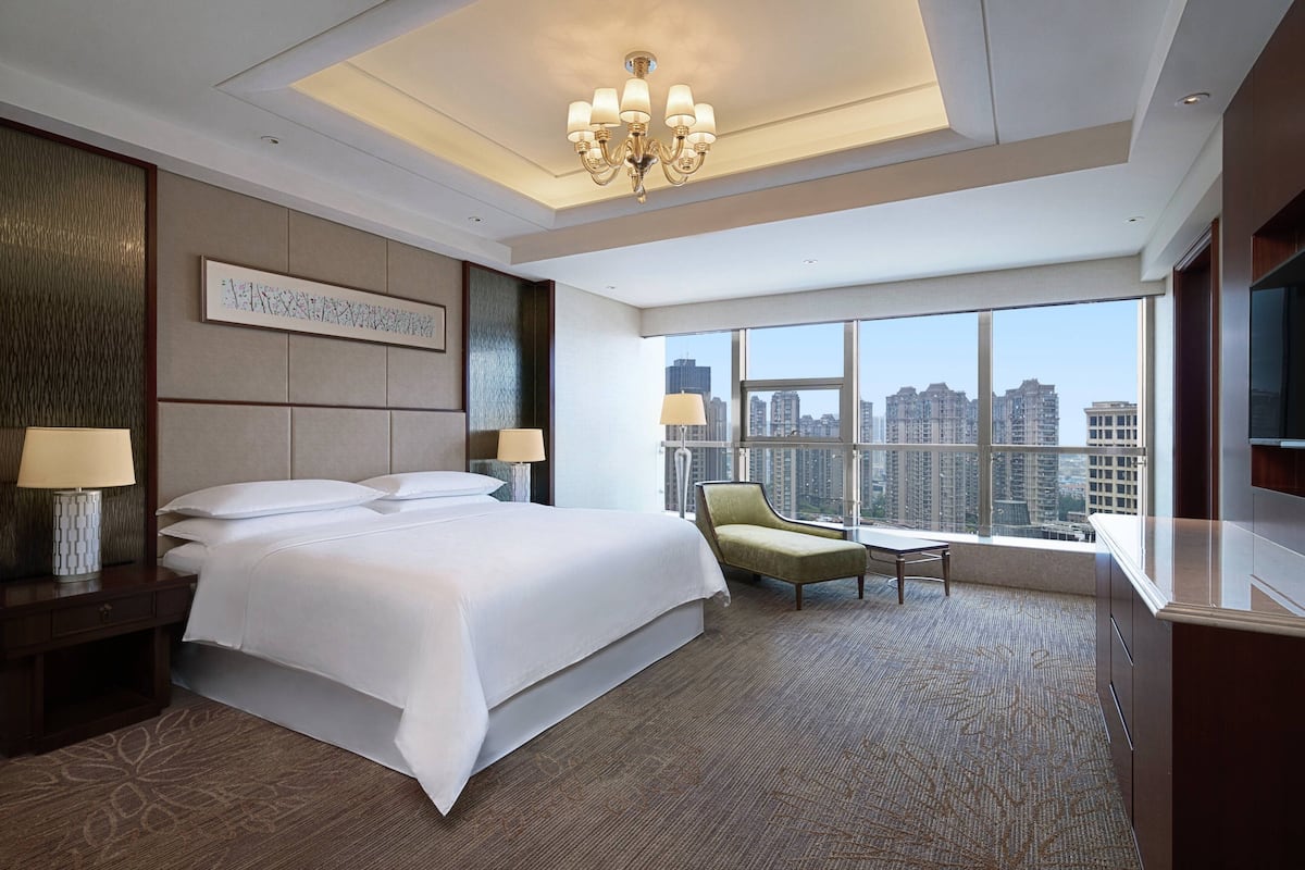 Una habitación de hotel moderna con una cama grande, una silla, un escritorio y vistas a la ciudad a través de amplias ventanas.