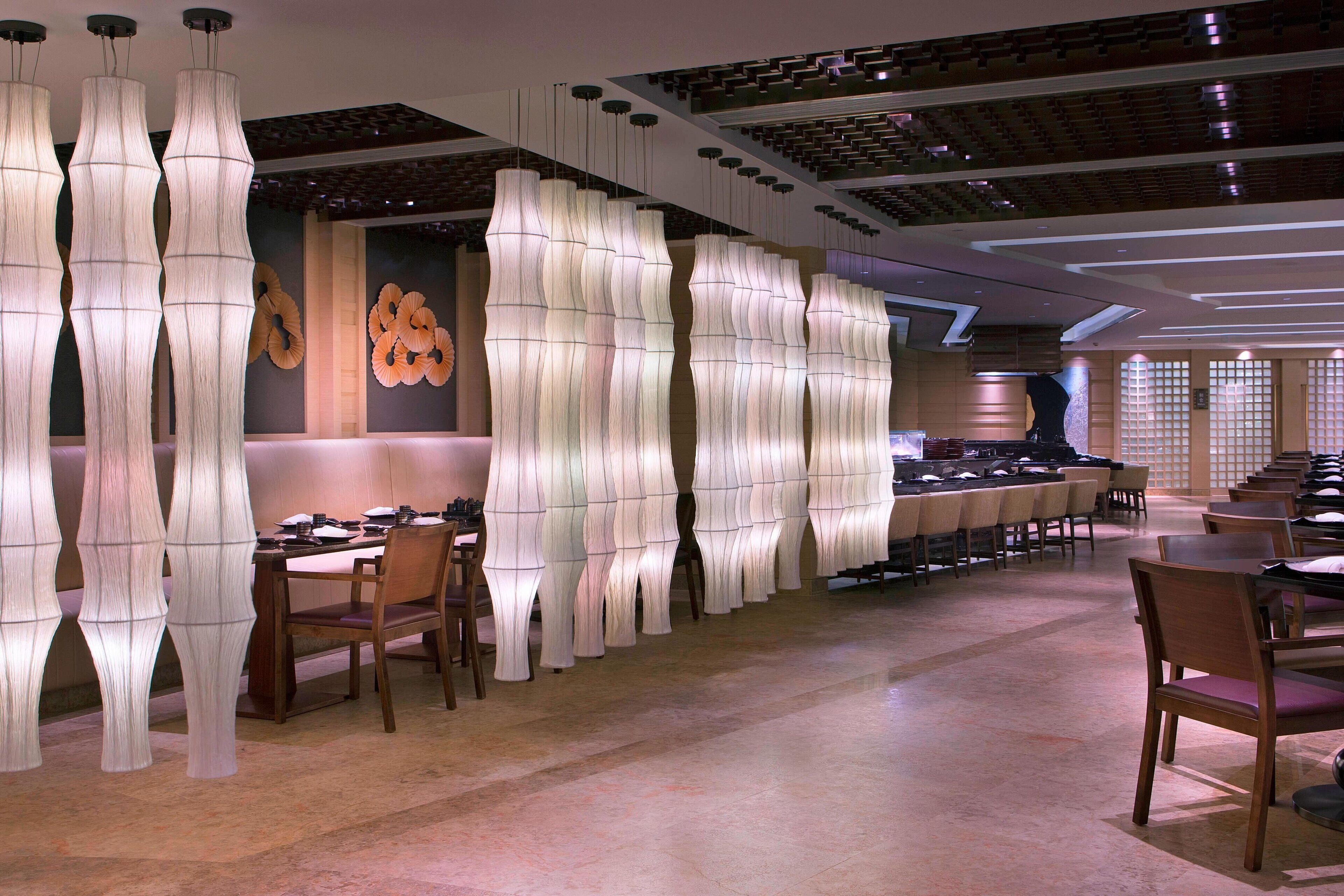 Un restaurante con decoración moderna, que presenta lámparas blancas y abstractas de gran altura y mesas y sillas de madera.