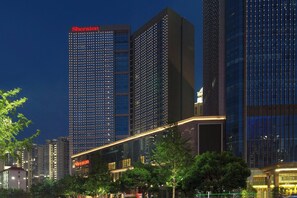 Un hotel moderno de gran altura con un llamativo letrero 'Sheraton', rodeado de otros edificios altos y árboles.