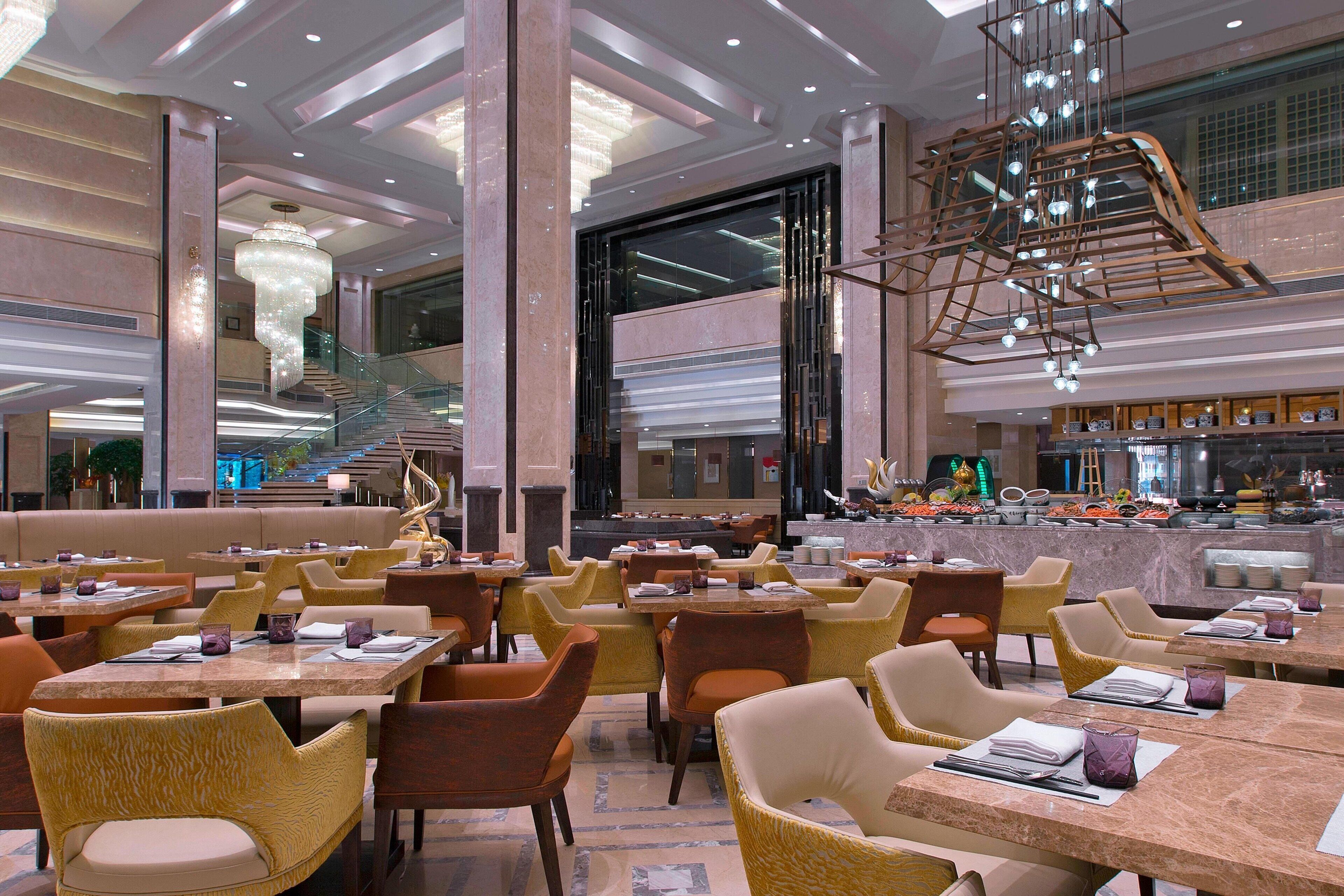 Un amplio vestíbulo de hotel con varias mesas y sillas de comedor, una escalera monumental y un moderno candelabro.