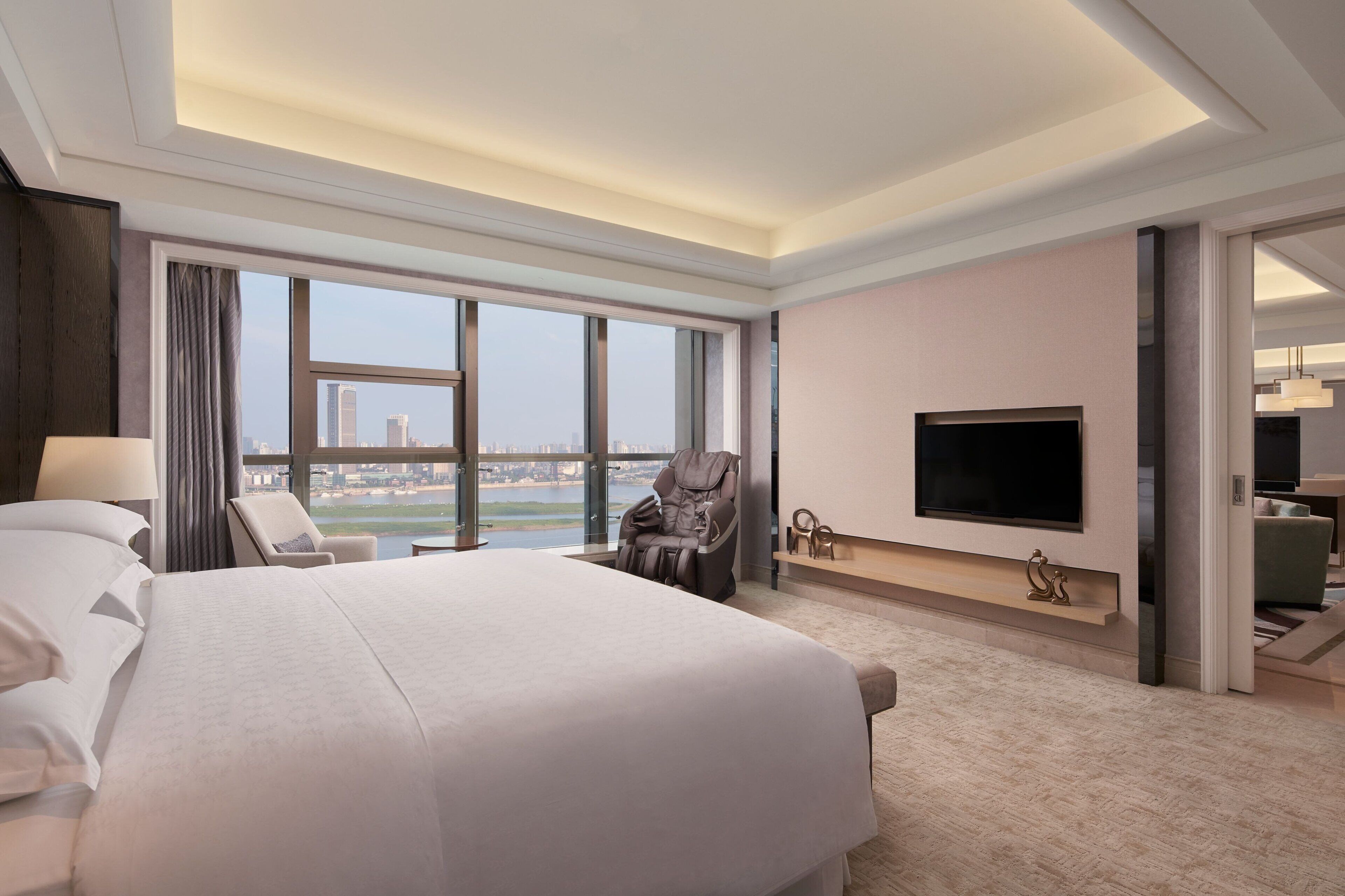 A hotel room with a large bed, a massage chair, a flat-screen TV, and a view of the cityscape.