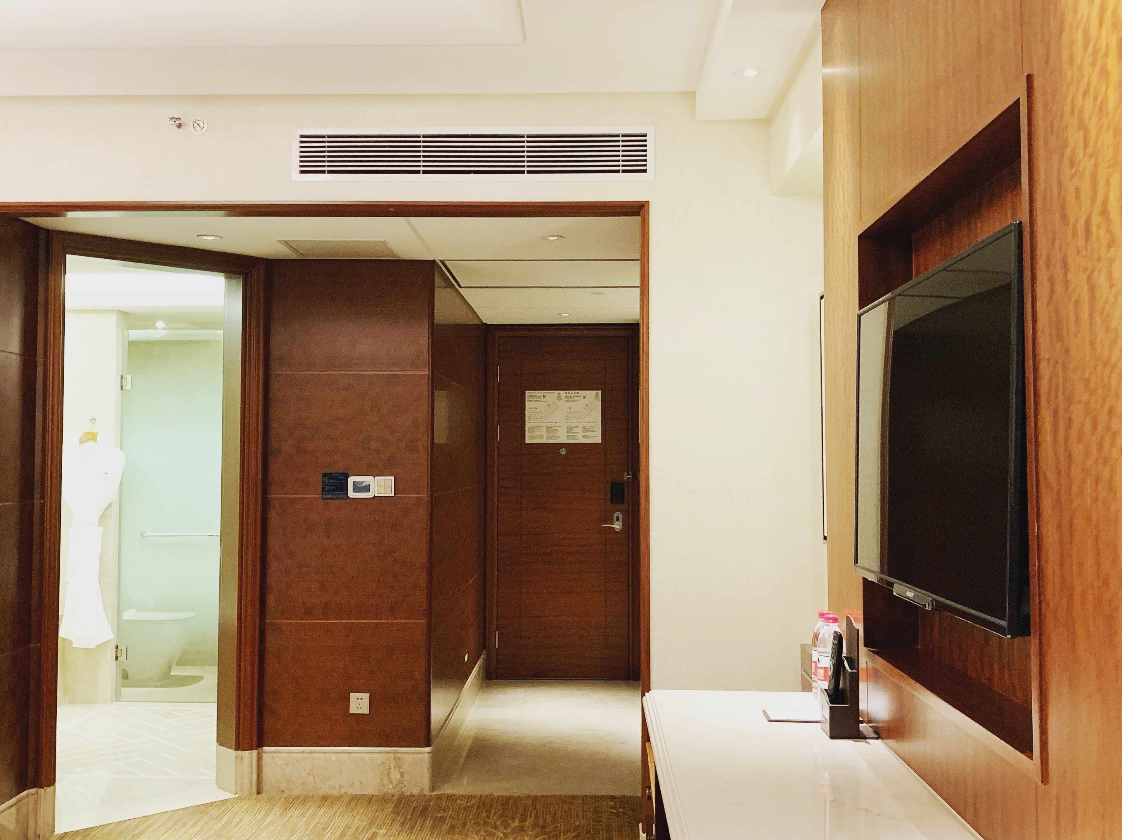 Un pasillo de hotel con una puerta de madera, una pared blanca, una rejilla de ventilación y un televisor fijado a la pared.